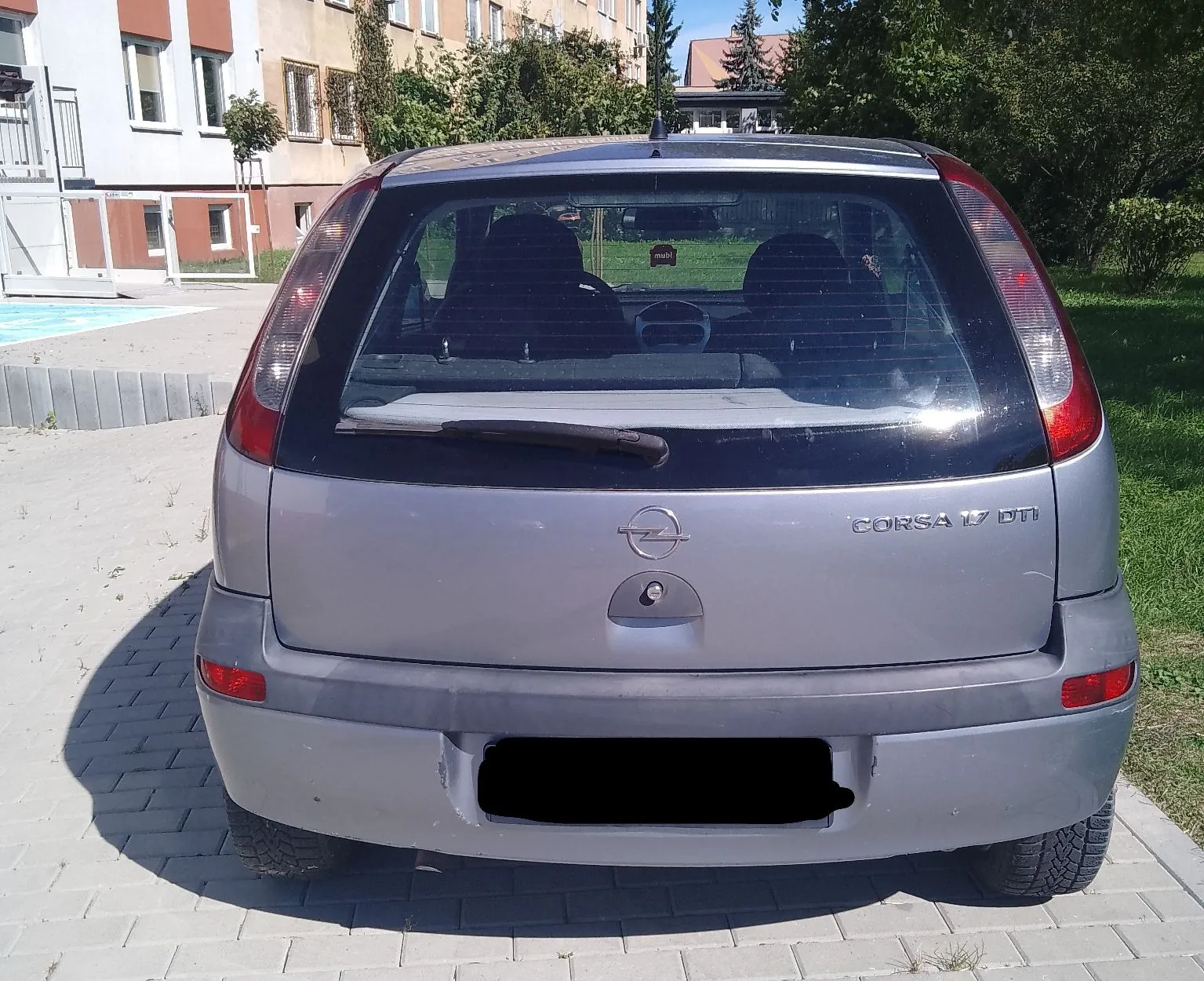 Tylny widok srebrnego samochodu marki Opel Corsa. Na klapie bagażnika znajduje się logo Opela oraz napis 'CORSA 1.7 DTI'. Samochód zaparkowany jest na kostce brukowej przed budynkiem mieszkalnym. Tablica rejestracyjna jest zakryta czarnym polem.