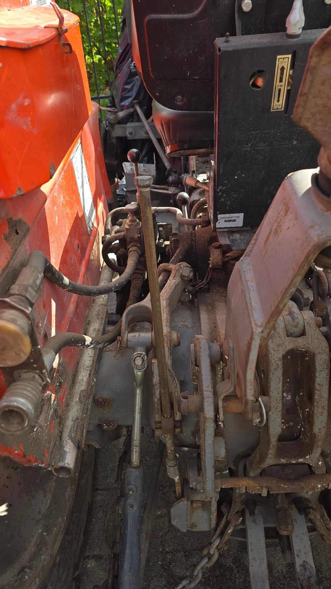 Zbliżenie na zardzewiałe mechanizmy i podzespoły techniczne ciągnika rolniczego. Po lewej stronie widoczny jest pomarańczowy element karoserii, a w centrum metalowe dźwignie, przewody hydrauliczne oraz masywne, metalowe części konstrukcyjne pokryte rdzą i brudem.