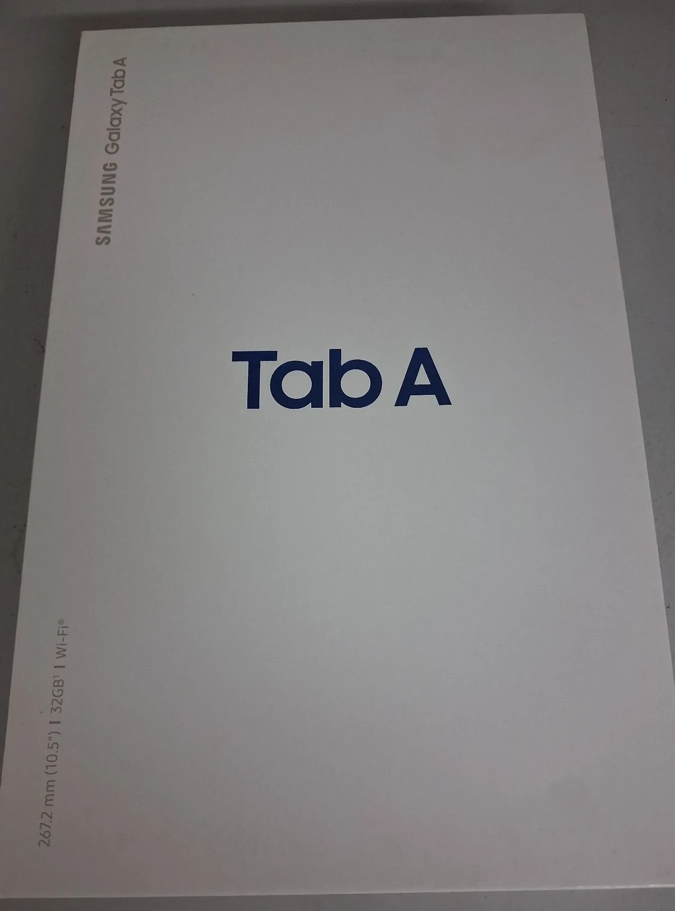 Białe pudełko tabletu z napisem 'Tab A' na środku. W górnej lewej części znajduje się pionowy napis 'SAMSUNG Galaxy Tab A', a w dolnej lewej części pionowy tekst '267.2 mm (10.5") | 32GB | Wi-Fi®'.