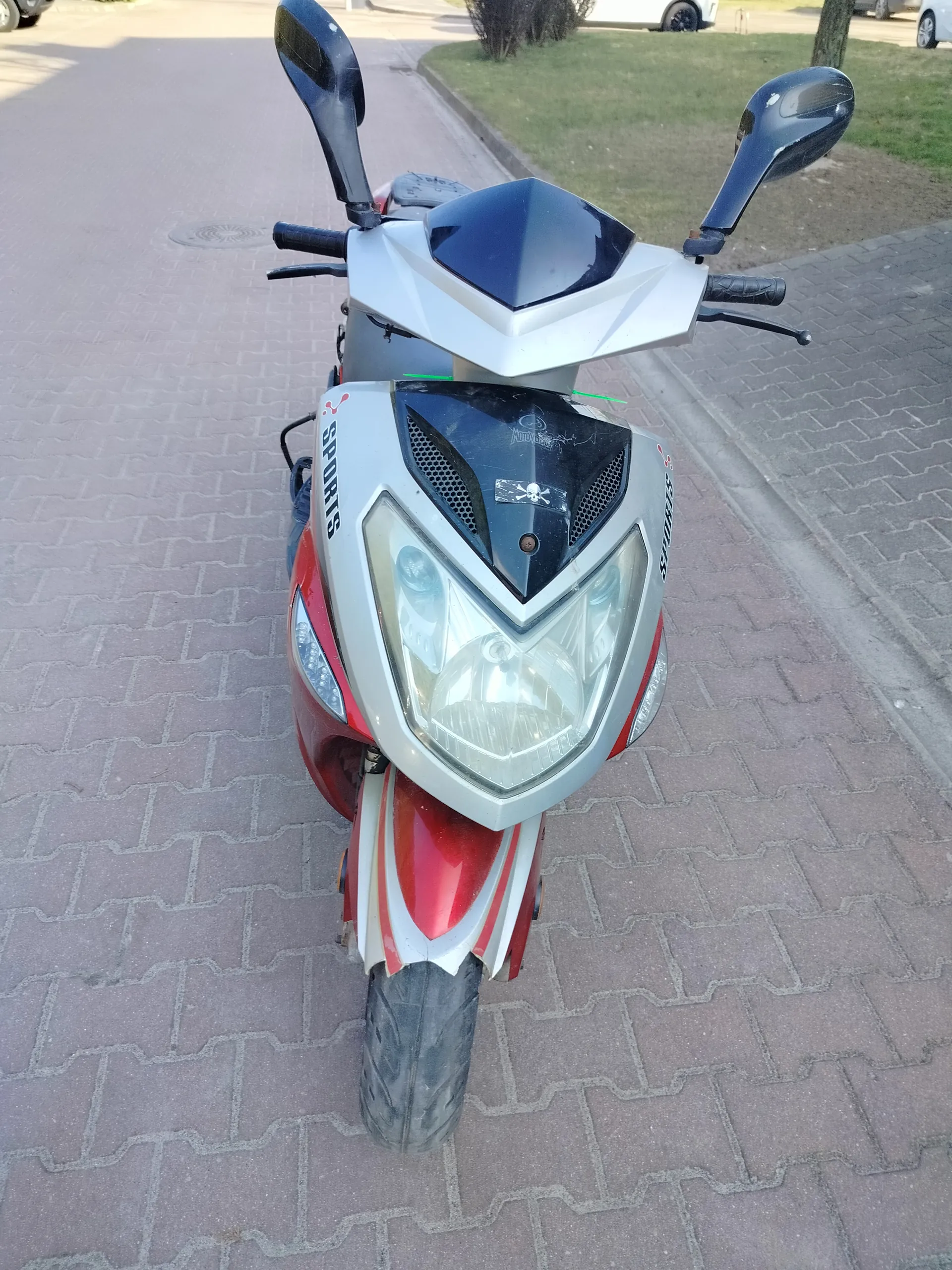 Czerwono-srebrny skuter widziany od przodu, z napisem SPORTS na bokach, dużym reflektorem, czarnym osłoną przednią z naklejką czaszki i krzyżyka, lusterkami bocznymi oraz widocznymi kierownicą i przednim kołem. Tło stanowi brukowana powierzchnia, trawnik i częściowo widoczne samochody zaparkowane w oddali.