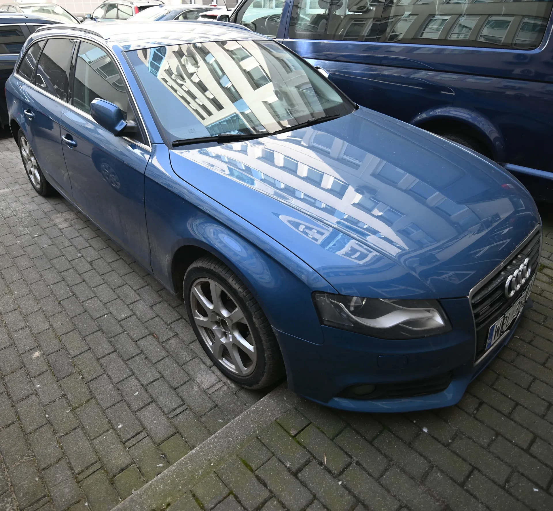 Niebieski samochód Audi typu kombi zaparkowany na brukowanej powierzchni. Widoczne są reflektory, tablica rejestracyjna, felgi oraz odbicie budynków w szybie przedniej. W tle znajdują się inne pojazdy, w tym niebieski samochód osobowy. Tło stanowi chodnik z kostki brukowej i fragmenty budynków odbijające się w szybach.