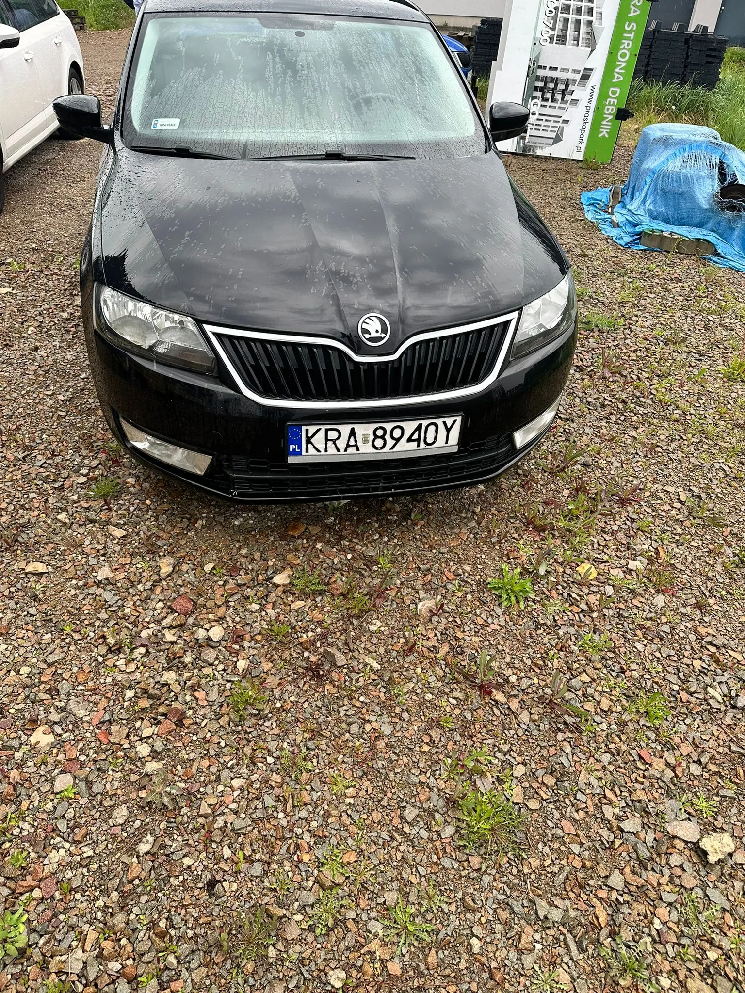 Czarny samochód marki Škoda z tablicą rejestracyjną KRA 8940Y (PL) zaparkowany na żwirowym podłożu. Na przedniej szybie widoczne krople deszczu. Po lewej stronie częściowo widoczny biały samochód, po prawej niebieska folia zakrywająca przedmiot oraz zielony znak z napisem 'STRONA DÉBNIK' i adresem www.parkpark.pl. Tło stanowi trawa i budynki.