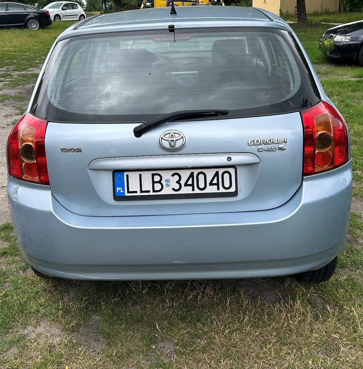 Tylny widok jasnoniebieskiego samochodu marki Toyota Corolla. Na klapie bagażnika widoczne jest logo Toyoty, napis 'TOYOTA' po lewej stronie oraz napis 'COROLLA' i mniejszy tekst poniżej po prawej stronie. Samochód posiada polską tablicę rejestracyjną o numerze LLB 34040. Pojazd stoi na trawiastym podłożu, w tle widoczne są inne samochody i drzewa.