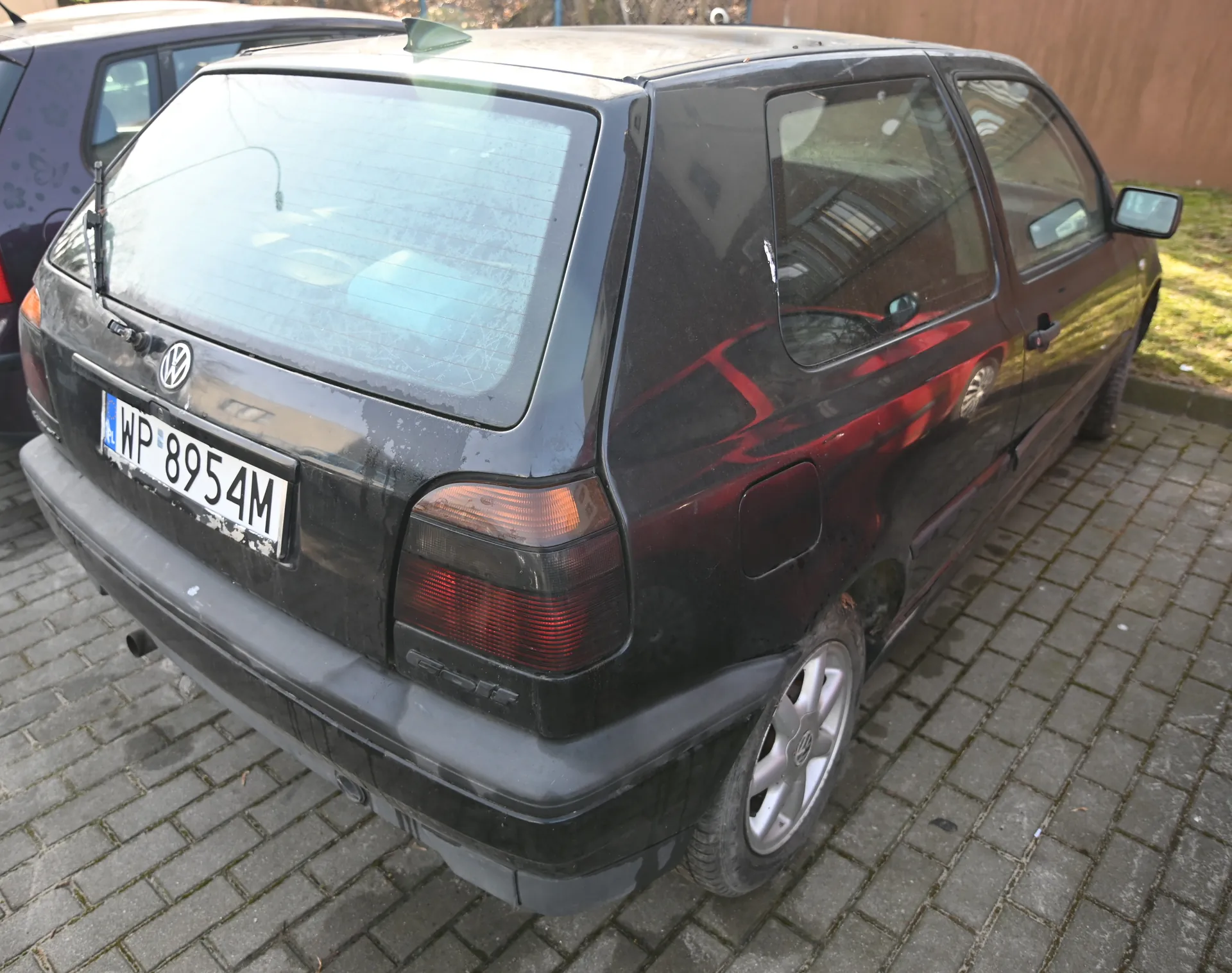 Czarny samochód Volkswagen Golf z tablicą rejestracyjną WP 8954M zaparkowany na brukowanej powierzchni. Widoczne są tylne światła, logo VW, częściowo zabrudzone tylne szyby oraz srebrne felgi. W tle fragment fioletowego samochodu z motywem na drzwiach, drewniany płot i trawa. Samochód znajduje się w pozycji trzydrzwiowej, widok z tyłu i boku.