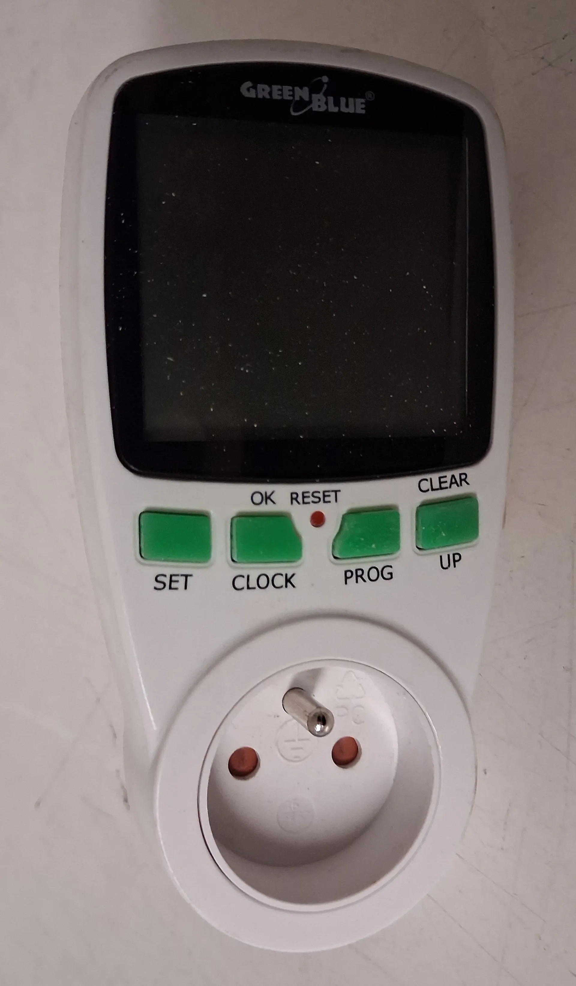 Białe urządzenie pomiarowe marki GREEN BLUE. W górnej części znajduje się czarny ekran LCD, a poniżej cztery zielone przyciski z podpisami: SET, CLOCK, PROG oraz CLEAR (z dodatkowym napisem UP). Nad przyciskami widnieją napisy OK i RESET oraz mała czerwona dioda. W dolnej części urządzenia znajduje się gniazdo elektryczne typu E z dwoma otworami na bolce i otworem uziemiającym.