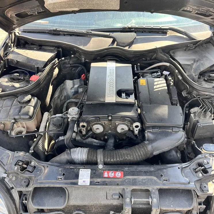 Widok z góry na komorę silnika czarnego samochodu marki Mercedes-Benz. W centrum znajduje się silnik z metalową osłoną z napisem 'Mercedes-Benz'. Widoczne są różne elementy mechaniczne, w tym rury dolotowe, zbiornik płynu chłodniczego po lewej stronie oraz plastikowe osłony. Obraz obejmuje fragment przedniej maski i przedniego pasa pojazdu.