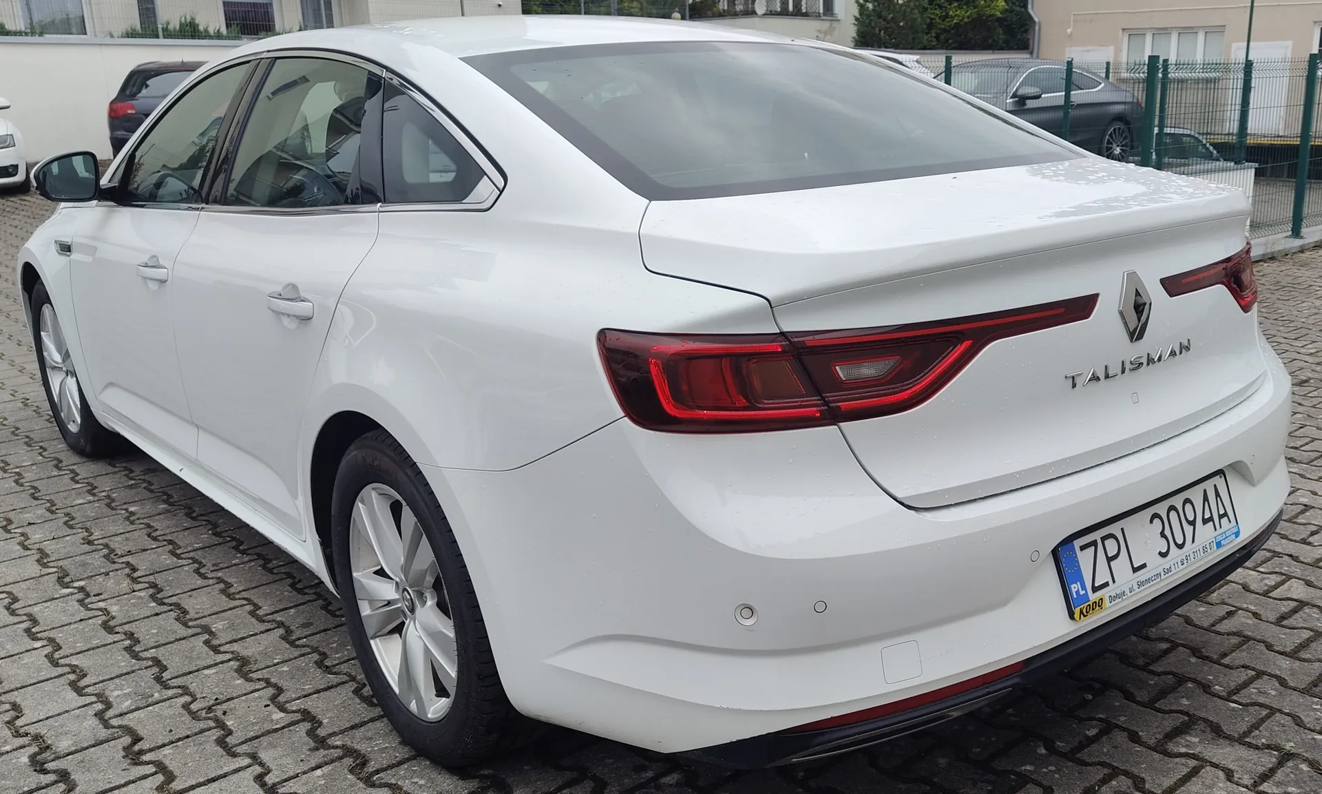 Biały samochód marki Renault Talisman zaparkowany na kostce brukowej. Widok z tyłu i z boku pojazdu. Na klapie bagażnika znajduje się logo Renault oraz napis 'TALISMAN'. Samochód ma polską tablicę rejestracyjną o numerze ZPL 3094A.