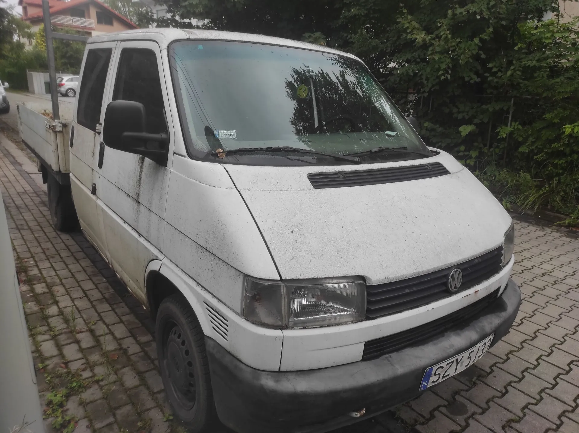 Biały samochód dostawczy marki Volkswagen z otwartą paką, zaparkowany na kostce brukowej. Pojazd jest zabrudzony, szczególnie w dolnych partiach nadwozia. Na przednim zderzaku widoczna jest tablica rejestracyjna o numerze SZY 5133. W tle znajdują się zielone drzewa i fragment budynku mieszkalnego.