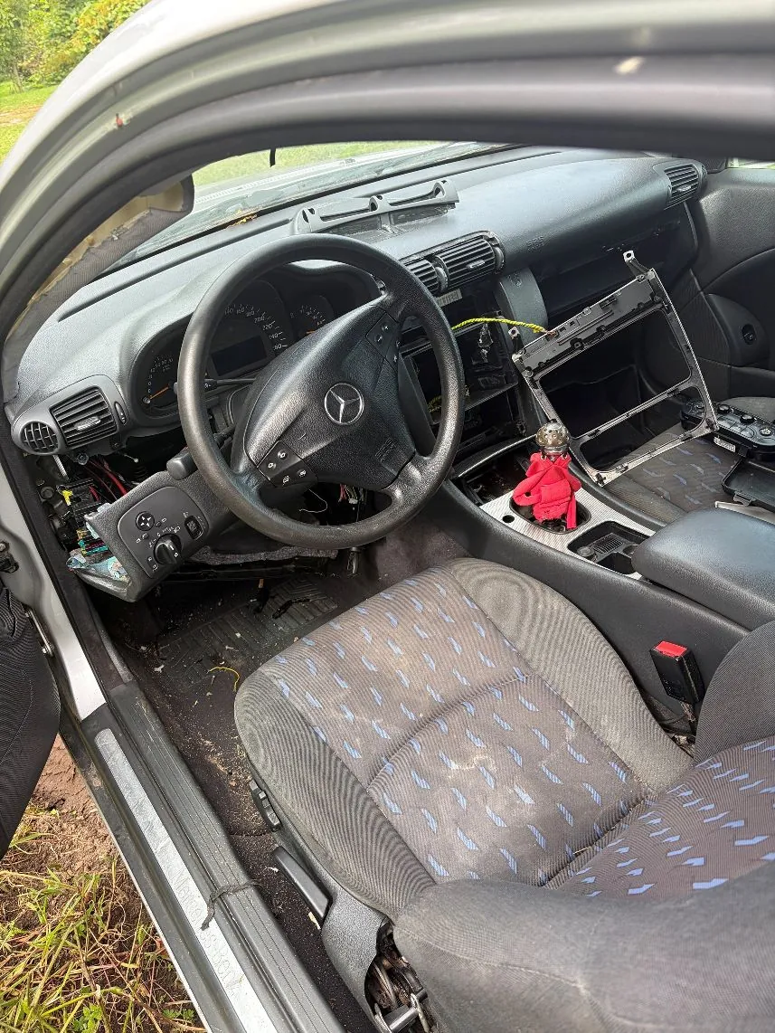 Wnętrze samochodu marki Mercedes-Benz z perspektywy otwartych drzwi kierowcy. Widoczna jest kierownica z logo marki, deska rozdzielcza z brakującymi elementami w centralnej części, gdzie odsłonięte są metalowe stelaże i kable. Na konsoli środkowej znajduje się mała czerwona figurka. Fotel kierowcy jest obity szarym materiałem w niebieskie wzory.