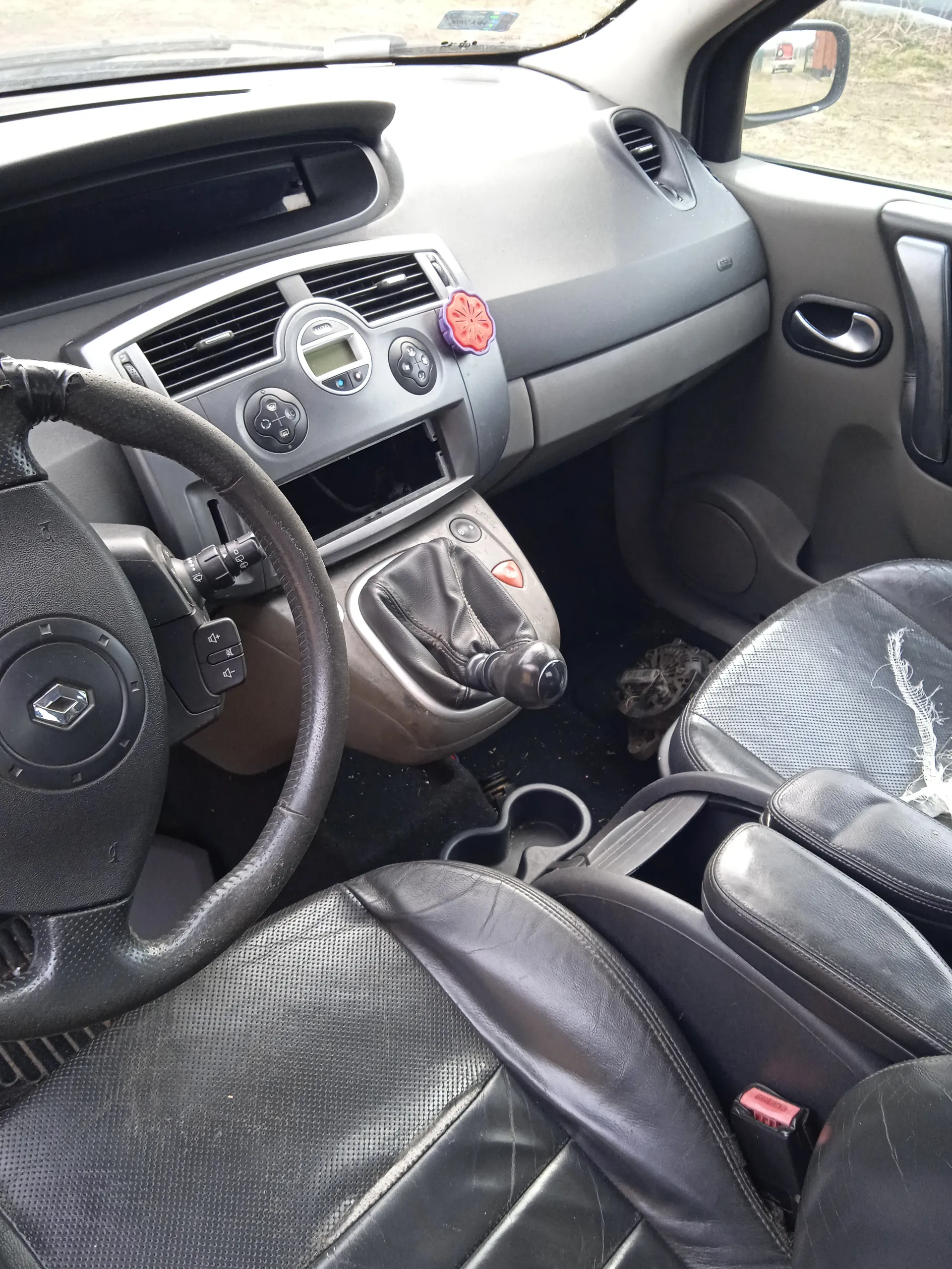 Wnętrze samochodu marki Renault. Widoczna kierownica z logo Renault, deska rozdzielcza z przyciskami i wyświetlaczem, dźwignia zmiany biegów z czarną osłoną, czarne skórzane fotele z widocznymi uszkodzeniami na siedzeniu pasażera. W tle przednia szyba, lusterko boczne oraz fragment zewnętrznej scenerii z trawą i drogą. Na desce rozdzielczej znajduje się czerwono-fioletowy naklejony element ozdobny.