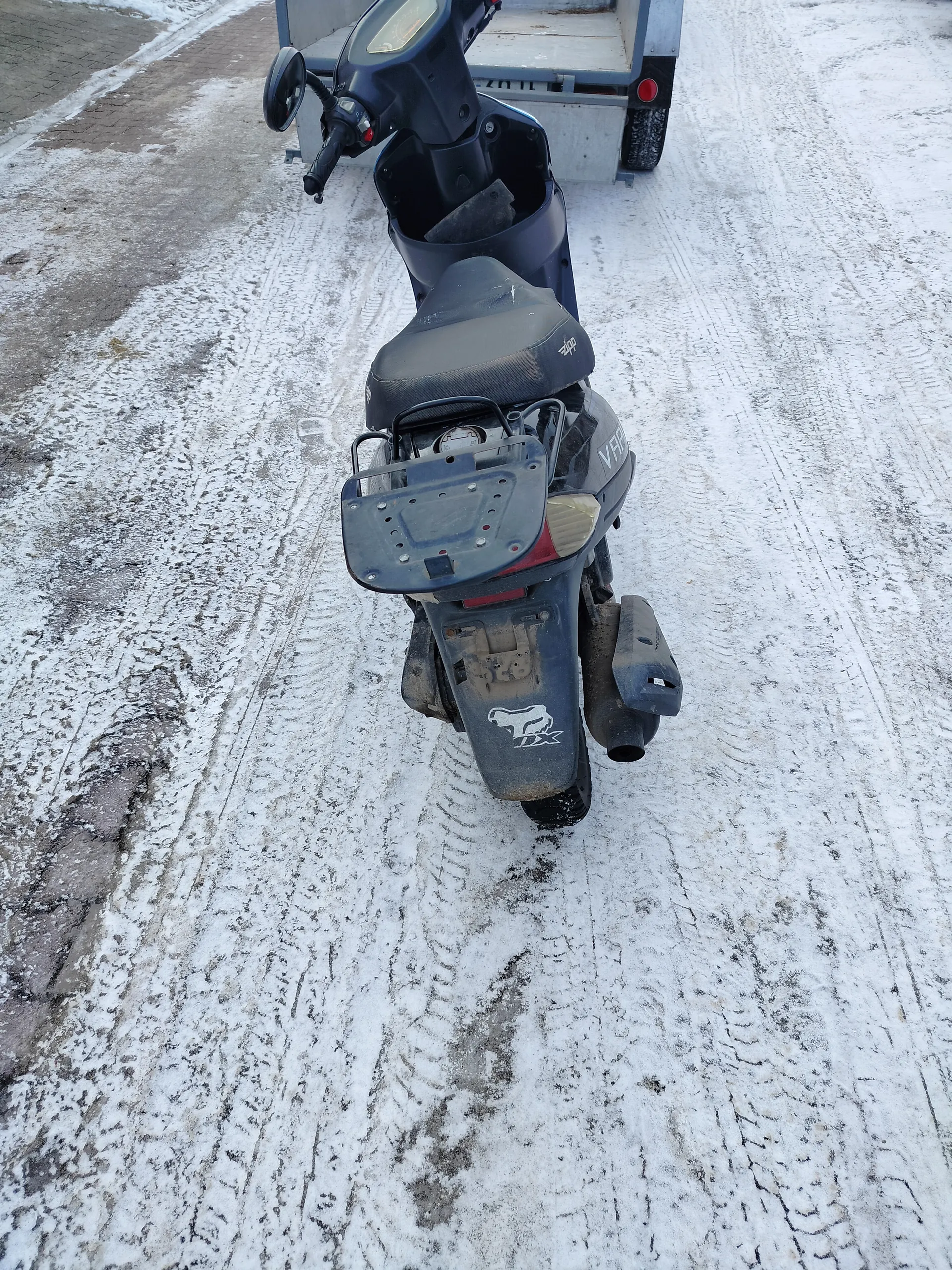 Czarny skuter zaparkowany na zasypanej śniegiem i lodem drodze z widocznymi śladami opon. Skuter posiada czarne siedzenie, lusterko w lewym kierunku oraz przyczepę z tyłu. Na tylnej części skutera znajduje się biały symbol zwierzęcia z napisem DX. Droga pokryta jest cienką warstwą śniegu z odsłoniętymi fragmentami asfaltu. W tle widoczna jest przyczepa z czerwonym światłem.