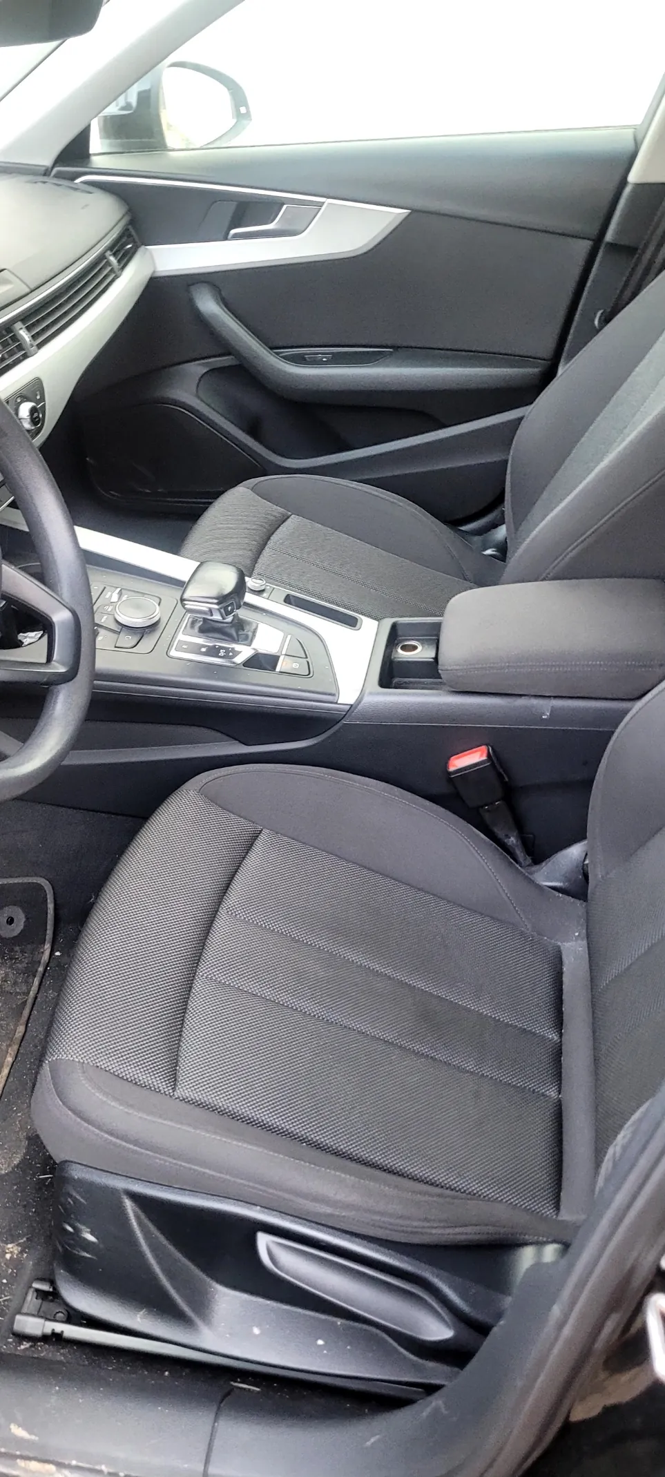 Wnętrze samochodu z czarnymi siedzeniami tapicerowanymi tkaniną, kierownicą po lewej stronie, centralną konsolą z dźwignią zmiany biegów, przyciskami sterowania i panelami drzwiowymi z uchwytem oraz sterowaniami okien. Widoczne są również pasy bezpieczeństwa i fragment deski rozdzielczej z wentylacją.