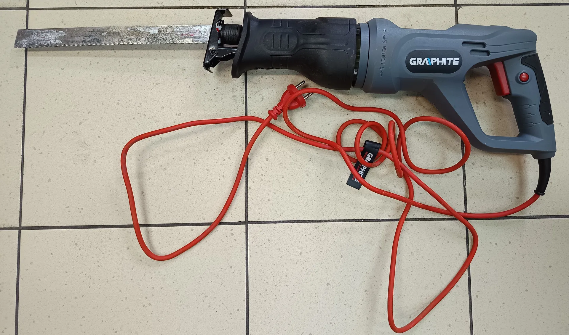 Recyprokacyjna piła marki GRAPHITE o szaro-czarnym kolorze z czerwonym przyciskiem i czerwonym przewodem zasilającym. Piła leży na jasnej płytkowej podłodze z ciemnymi szwami. Widoczna jest zużyta metalowa piła oraz czarna etykieta z napisem GRAPHITE. Na korpusie narzędzia znajduje się napis GRAPHITE oraz dodatkowe oznaczenia.