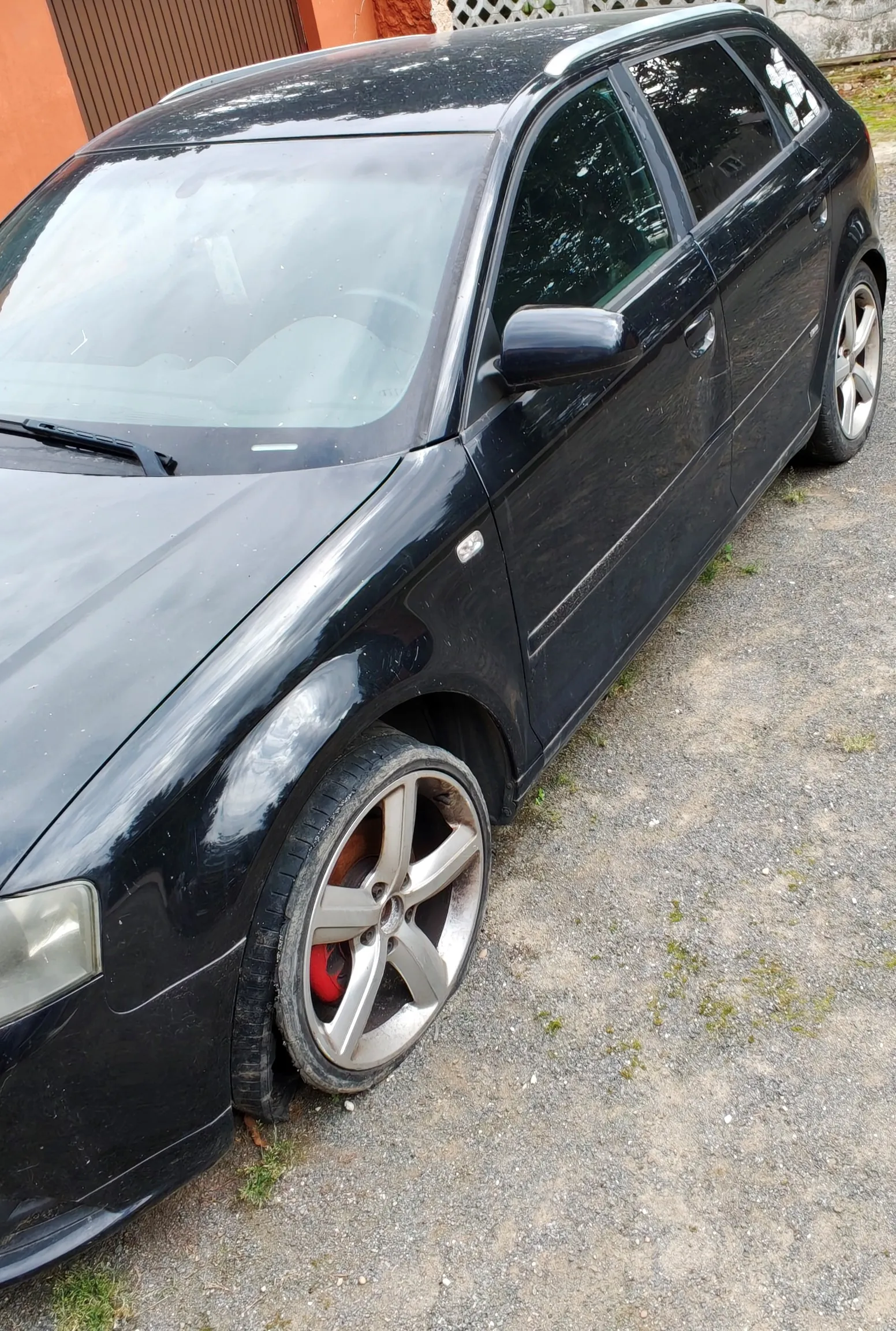 Czarny samochód typu kombi zaparkowany na żwirowym podjeździe. Zdjęcie przedstawione z perspektywy przedniego lewego błotnika i koła. Samochód ma srebrne pięcioramienne felgi z widocznymi czerwonymi zaciskami hamulcowymi.