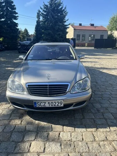 Srebrny samochód marki Mercedes-Benz zaparkowany przodem do obiektywu na brukowanym placu. Pojazd posiada polską tablicę rejestracyjną o numerze SCZ 10229. W tle widoczne są wysokie drzewa iglaste oraz jasny, parterowy budynek z ciemnym dachem.