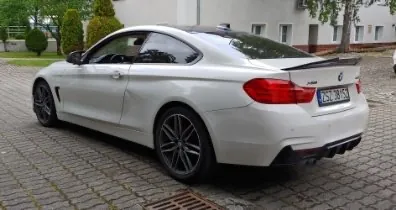 Biały samochód osobowy marki BMW w nadwoziu coupe, zaparkowany tyłem i bokiem na kostce brukowej. Pojazd posiada ciemne felgi aluminiowe, czarny spojler na klapie bagażnika oraz czarny dyfuzor tylnego zderzaka. Widoczna jest tablica rejestracyjna o numerze ZSZ 381SZ.
