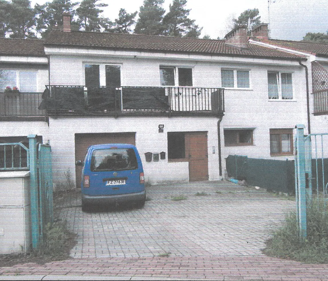 Biały dom dwupiętrowy z brązowym dachem krytym dachówką. Na pierwszym piętrze balkon z czarnym ogrodzeniem. Przy budynku niebieski samochód osobowy z tablicą rejestracyjną FZ 2763E. Widoczne są brama garażowa, brązowe drzwi wejściowe, okna oraz zielone ogrodzenie po prawej stronie. Dziedziniec wyłożony ceglaną kostką, na tle drzewa. Tekst na tablicy rejestracyjnej: FZ 2763E.