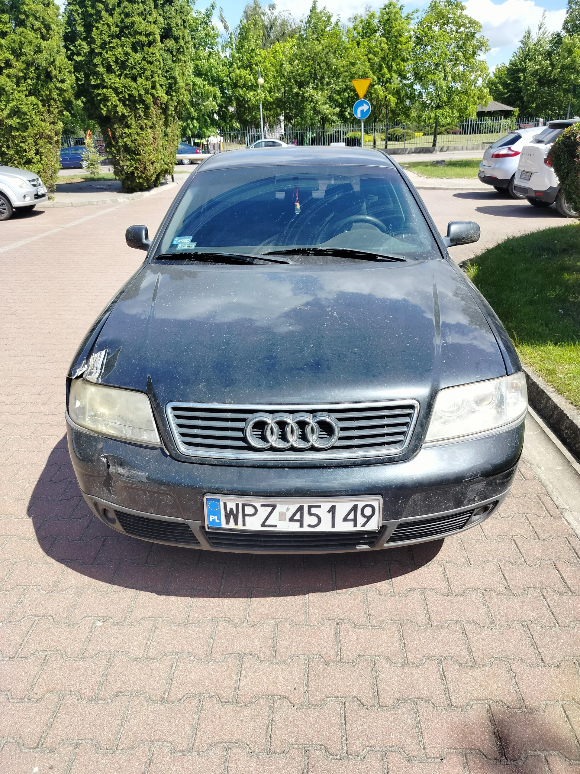 Czarny samochód marki Audi zaparkowany na brukowanej powierzchni. Widoczna tablica rejestracyjna z polskim kodem PL i numerem WPZ 45149. Przednia część pojazdu ma widoczne uszkodzenia po lewej stronie. W tle drzewa, inne zaparkowane samochody, znaki drogowe (żółty trójkąt i niebieski okrąg z białą strzałką w prawo) oraz ogrodzenie. Niebo częściowo zachmurzone.