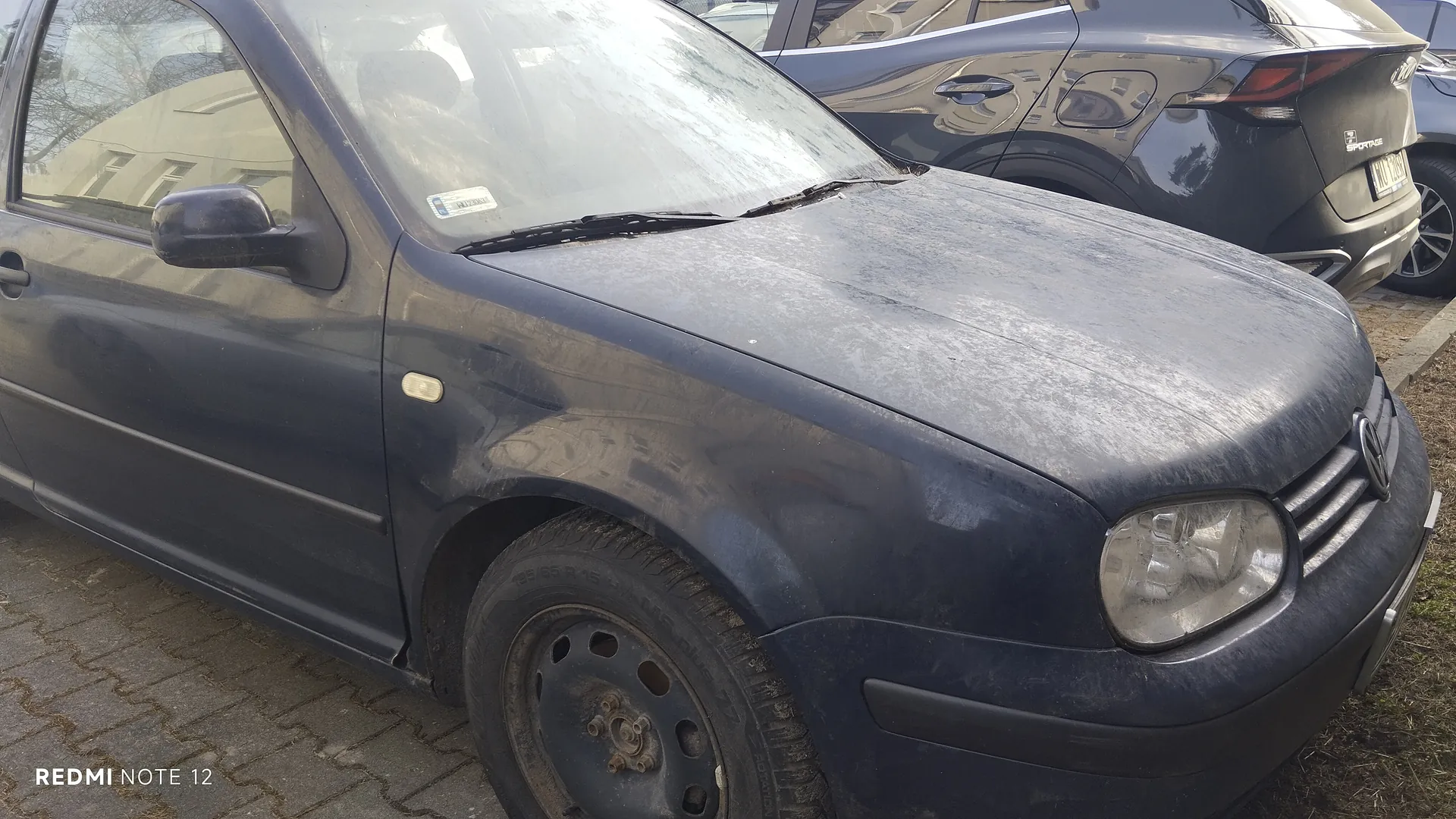 Zdjęcie przedstawia ciemnoniebieski samochód marki Volkswagen (model Golf) zaparkowany na brukowanej powierzchni. Samochód jest zabrudzony, widoczna kurz na masce i oponie. W tle znajduje się szary samochód z częściowo widoczną tablicą rejestracyjną. Na lewym dolnym rogu zdjęcia widnieje napis 'REDMI NOTE 12'.