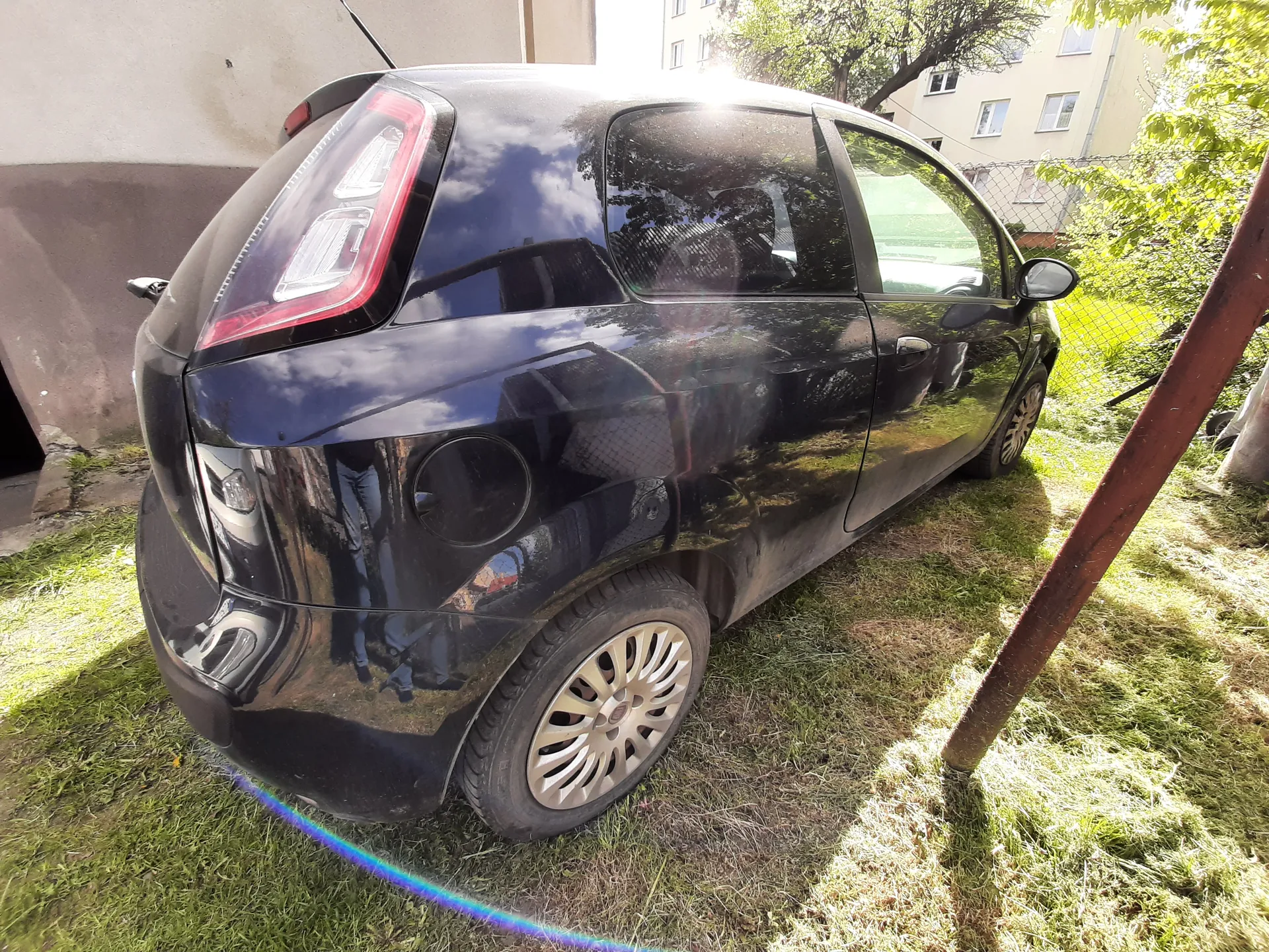 Czarny samochód osobowy typu hatchback zaparkowany na trawie. Widok z tyłu i boku. Pojazd ma błyszczącą karoserię odbijającą niebo, chmury i otoczenie. W tle widać fragment budynku, siatkę ogrodzeniową, drzewa oraz inny budynek z oknami. Trawa jest częściowo wyschnięta, a na obrazie widoczne są refleksy słońca.