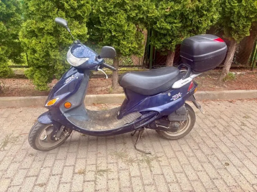 Niebieski skuter marki Kymco z zamontowanym czarnym kufrem z tyłu, zaparkowany na kostce brukowej na tle zielonych krzewów.