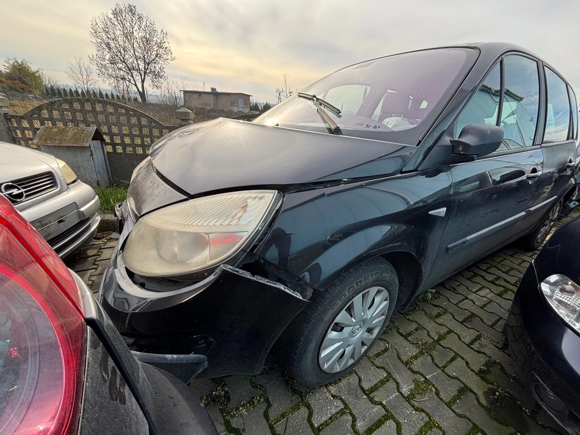 Zniszczony czarny samochód osobowy z uszkodzonym przodem, w tym pękniętą maską, zdeformowanym zderzakiem i uszkodzonym reflektorem. Samochód stoi na brukowanej powierzchni, otoczony innymi pojazdami: srebrnym po lewej stronie i częściowo widocznym czarnym po prawej. W tle widoczne są drewniane ogrodzenie, mały budynek, drzewo bez liści, dom oraz chmurne niebo. Na oponie widoczny napis 'GOODYEAR'.