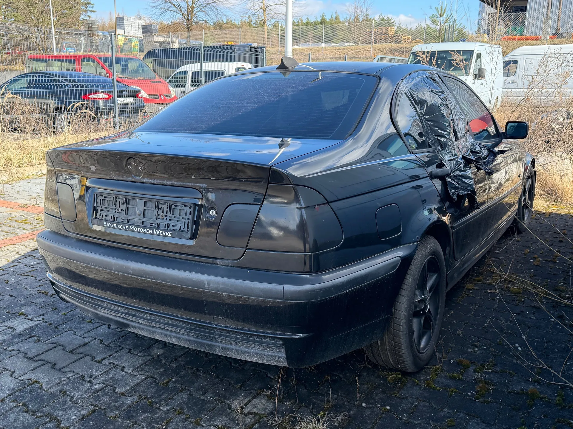 Czarny samochód osobowy typu sedan zaparkowany na brukowanej powierzchni. Widok z tyłu i boku. Na tylnej szybie widoczna folia ochronna. Tablica rejestracyjna z napisem "Bayerische Motoren Werke". W tle inne pojazdy (czerwony samochód, białe furgony), ogrodzenie metalowe, suche trawy i drzewa. Niebo jest jasne z kilkoma chmurami.