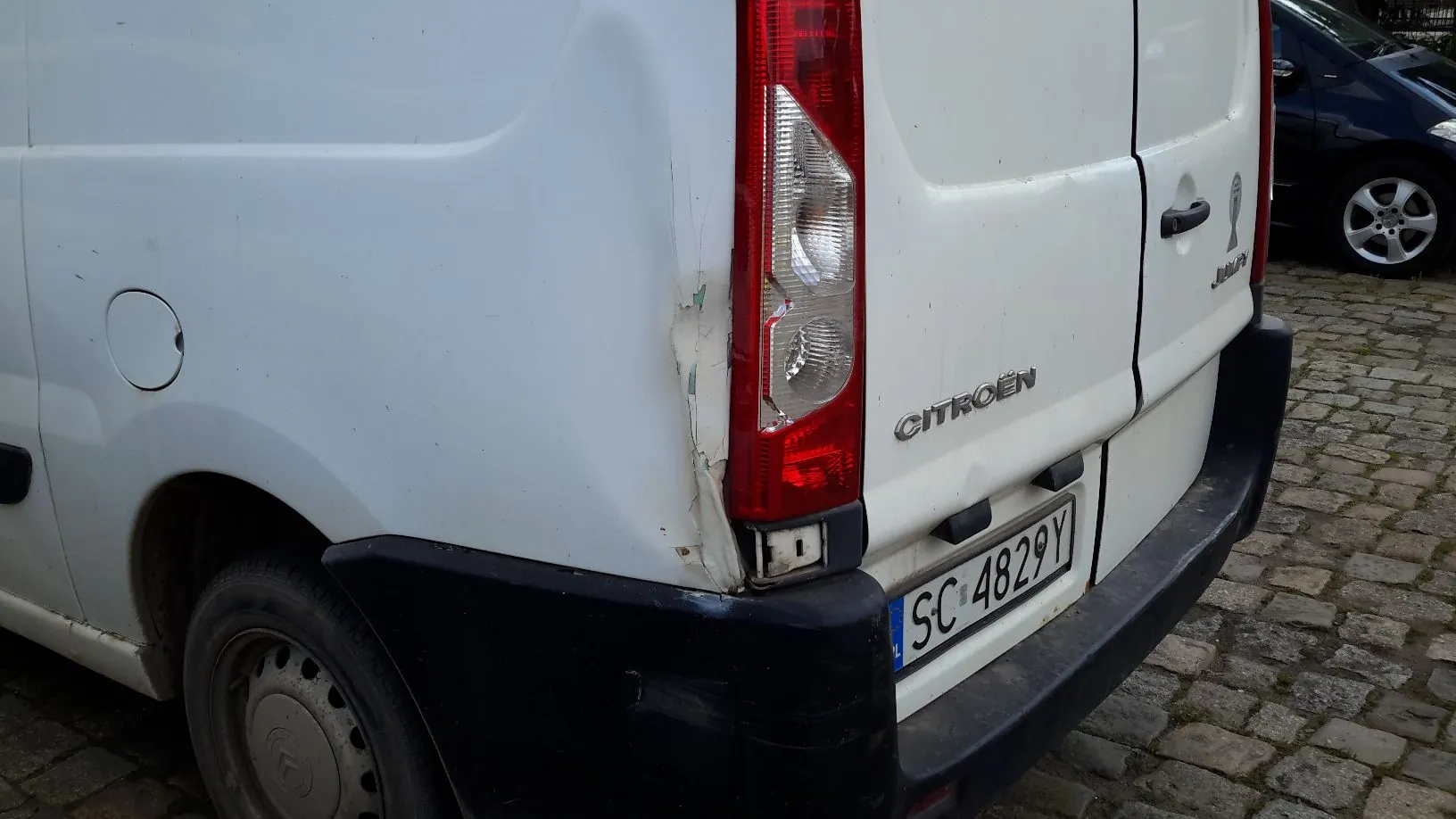 Zbliżenie na tył białego samochodu marki Citroën. Widoczna jest prawa tylna lampa, napis CITROËN oraz tablica rejestracyjna o numerze SC 4829Y. Na karoserii, obok lampy, widoczne są uszkodzenia w postaci wgnieceń i odprysków lakieru. Samochód stoi na kostce brukowej.