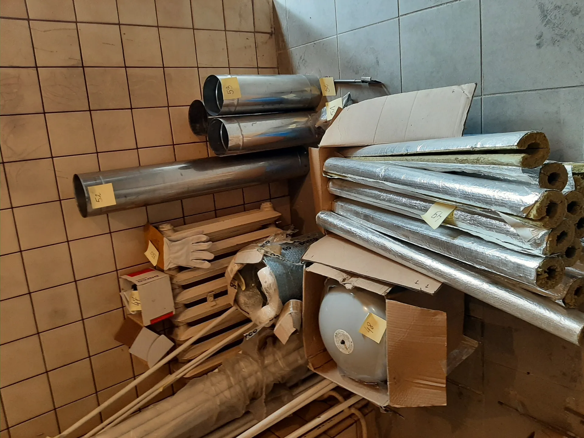 Zdjęcie przedstawia pomieszczenie magazynowe z różnymi przedmiotami. Po lewej stronie widoczna jest ściana wyłożona brązowymi kwadratowymi płytkami, po prawej – szarymi płytkami. W centrum znajdują się metalowe rury o różnych średnicach, niektóre z żółtymi etykietami z napisami: 52, 53, 67, 48. Obok rur leżą zwinięte materiały izolacyjne owinięte srebrną folią, częściowo w kartonowych pudełkach. W jednym z pudeł znajduje się okrągły, szary przedmiot z etykietą 48. W tle widoczne są białe rękawice, drewniane listwy oraz rozrzucone pudełka i opakowania. Tekst na etykietach jest czytelny i ustawiony pionowo.