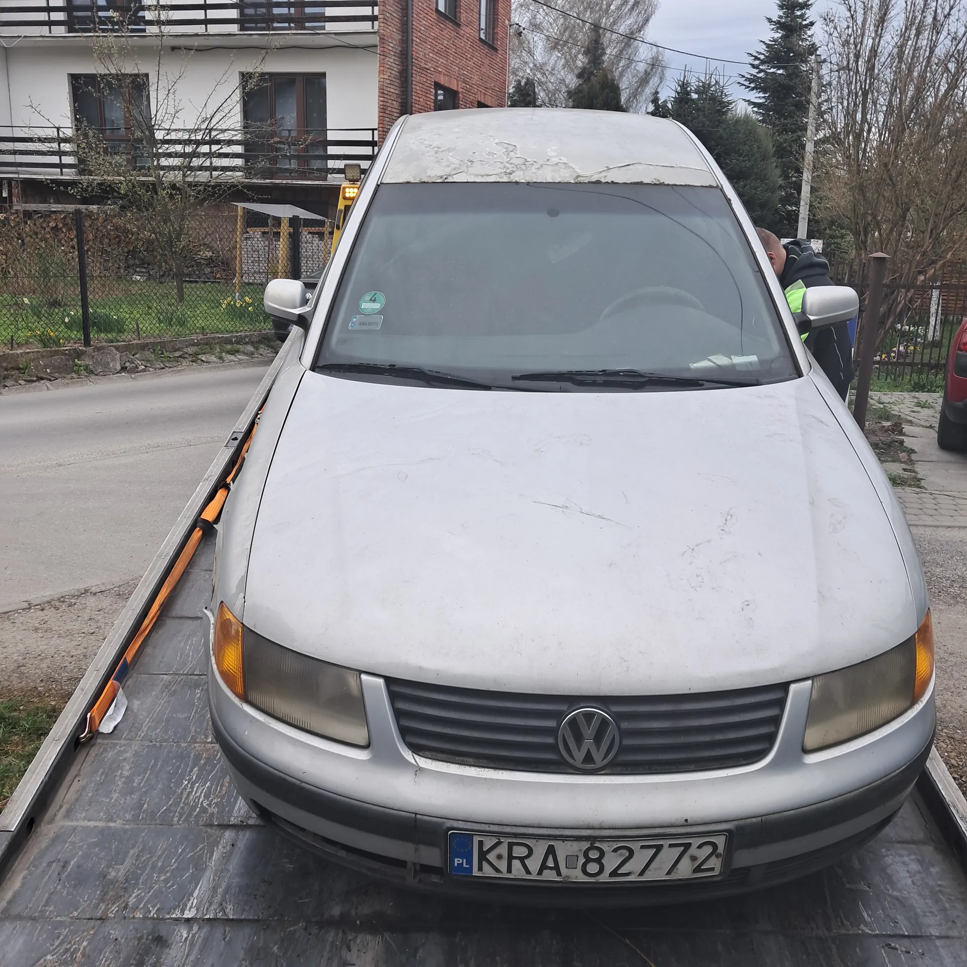 Srebrny samochód marki Volkswagen zaparkowany na poboczu. Widoczna tablica rejestracyjna z napisem KRA 82772 oraz oznaczeniem PL. Na masce samochodu znajdują się zadrapania. Przy kierowcy stoi osoba w żółto-zielonej kamizelce odblaskowej. W tle budynki mieszkalne, drzewa, ogrodzenie i droga.