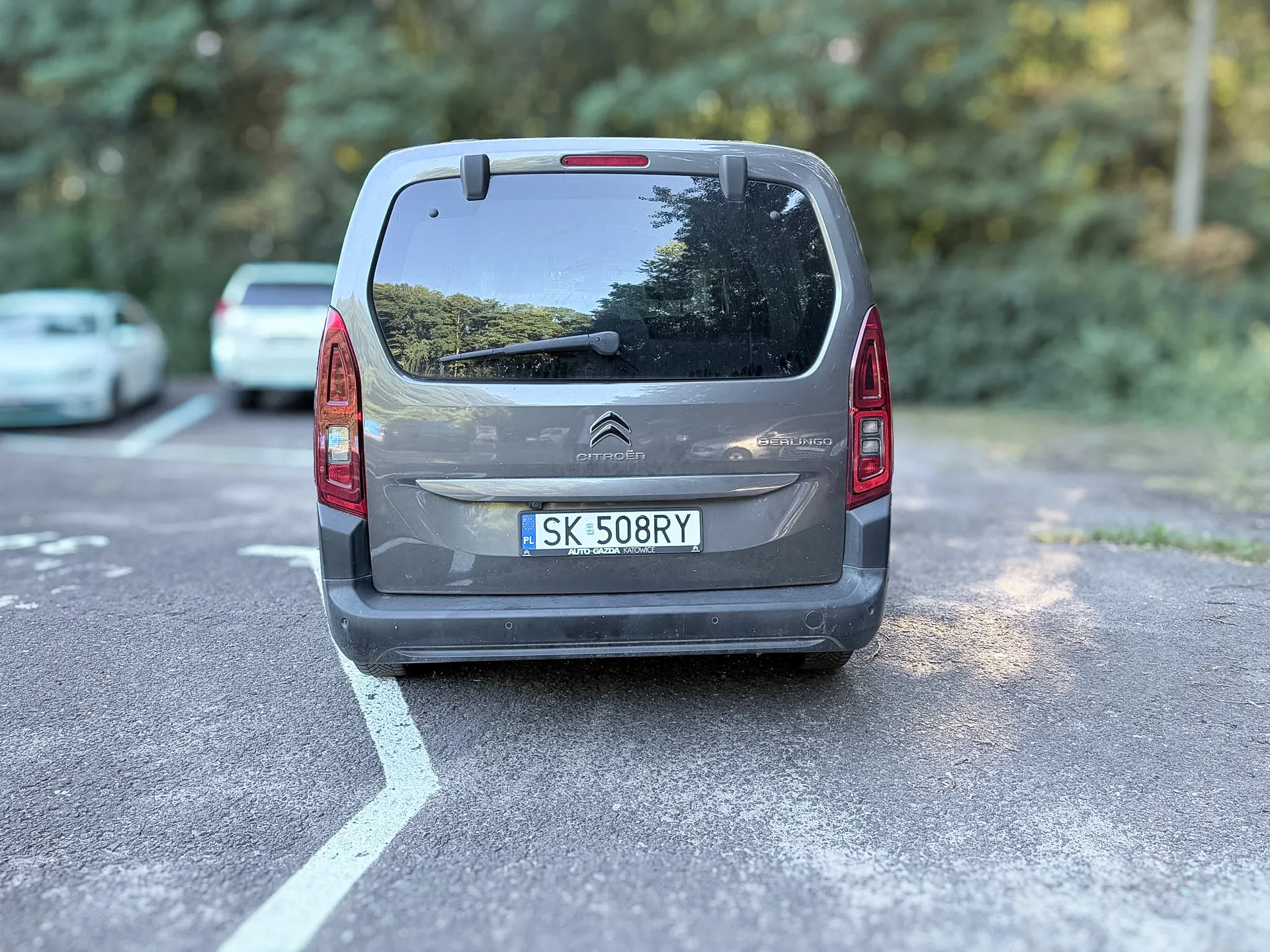 Tylny widok szarego samochodu marki Citroën Berlingo zaparkowanego na asfaltowym placu. Pojazd posiada polską tablicę rejestracyjną o numerze SK 508RY. W tle widoczne są inne samochody oraz zielone drzewa i krzewy.