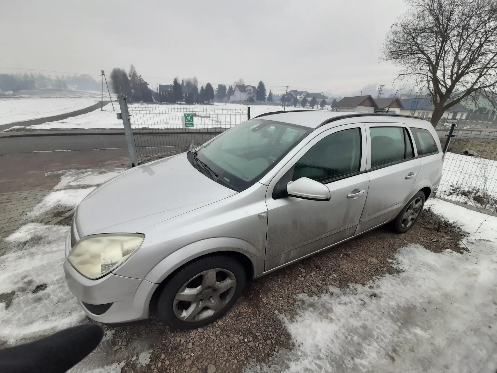 Srebrny samochód typu kombi zaparkowany na żwirowym obszarze z resztkami śniegu. W tle widoczna droga, metalowe ogrodzenie, domy, drzewa bez liści oraz zimowy krajobraz z śniegiem na polach. Niebo jest zachmurzone, a na ziemi znajdują się plamy śniegu i odsłoniętej ziemi. Samochód ma zabrudzone boki i przednią część.