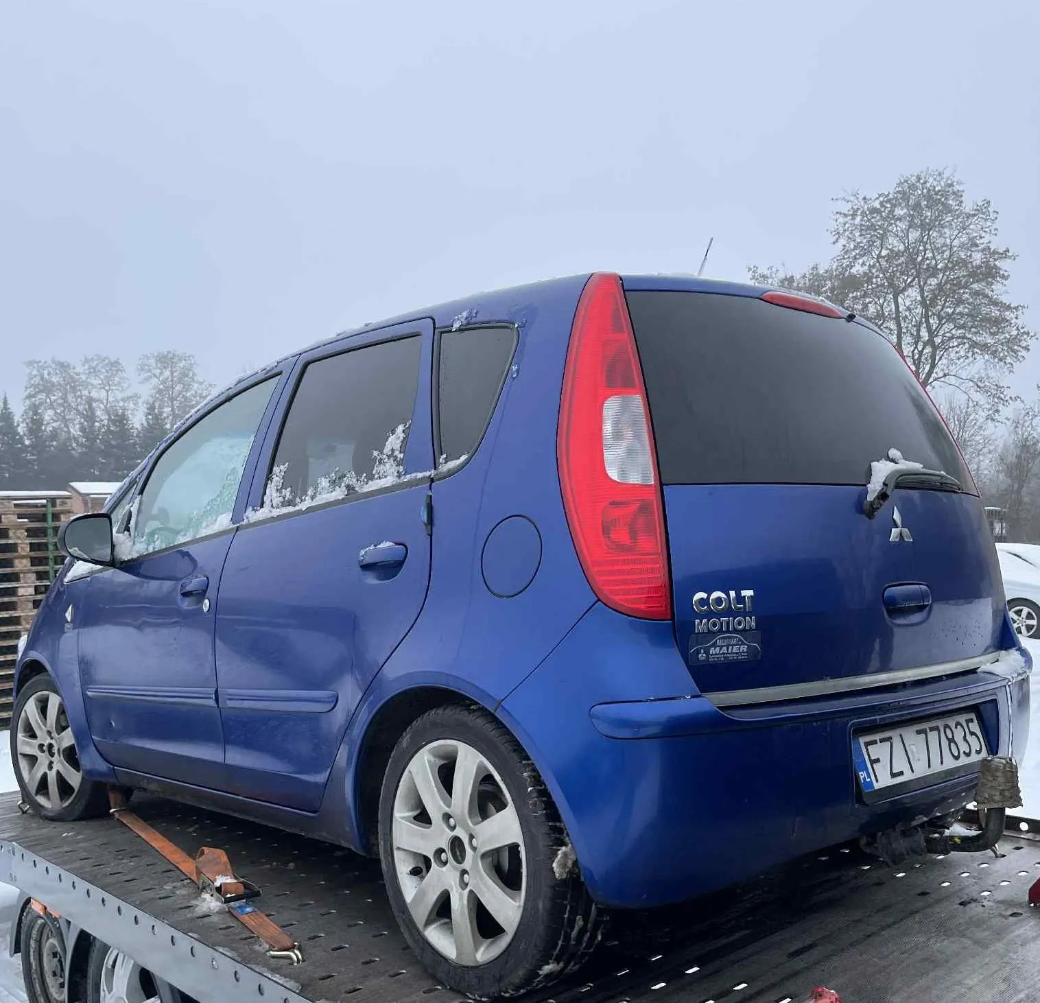 Niebieski samochód Mitsubishi Colt Motion z tablicą rejestracyjną FZ1 77835 (PL) umieszczony na płycie przyczepy. Na tylnym zderzaku widoczny napis COLT MOTION oraz naklejka MAIER. Samochód pokryty jest śniegiem, szczególnie na szybach i dachu. Tło przedstawia zalesiony teren w zimowej pogodzie z widocznymi drzewami i pochmurnym niebem. Na przyczepie znajdują się pomarańczowe paski mocujące.