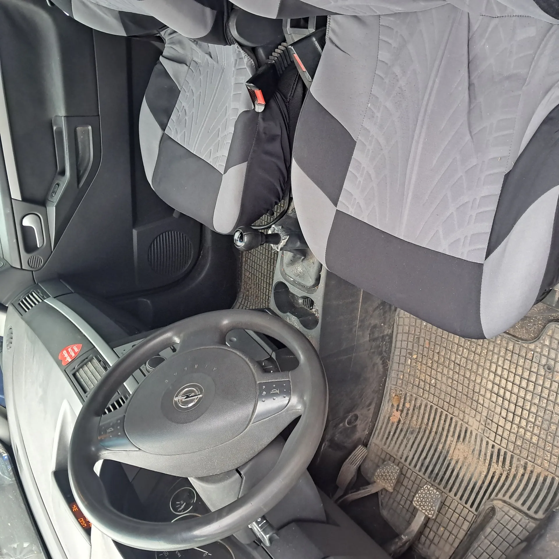 Wnętrze samochodu z widocznym kierownicą z logo Opel, siedzeniami w kolorze szaro-czarnym z geometrycznym wzorem, przyciskiem bezpieczeństwa z czerwonym elementem, pedałami, brudnymi dywanikami podłogowymi, deska rozdzielcza z czerwonym naklejką oraz panel drzwiowy z uchwytem i głośnikiem.