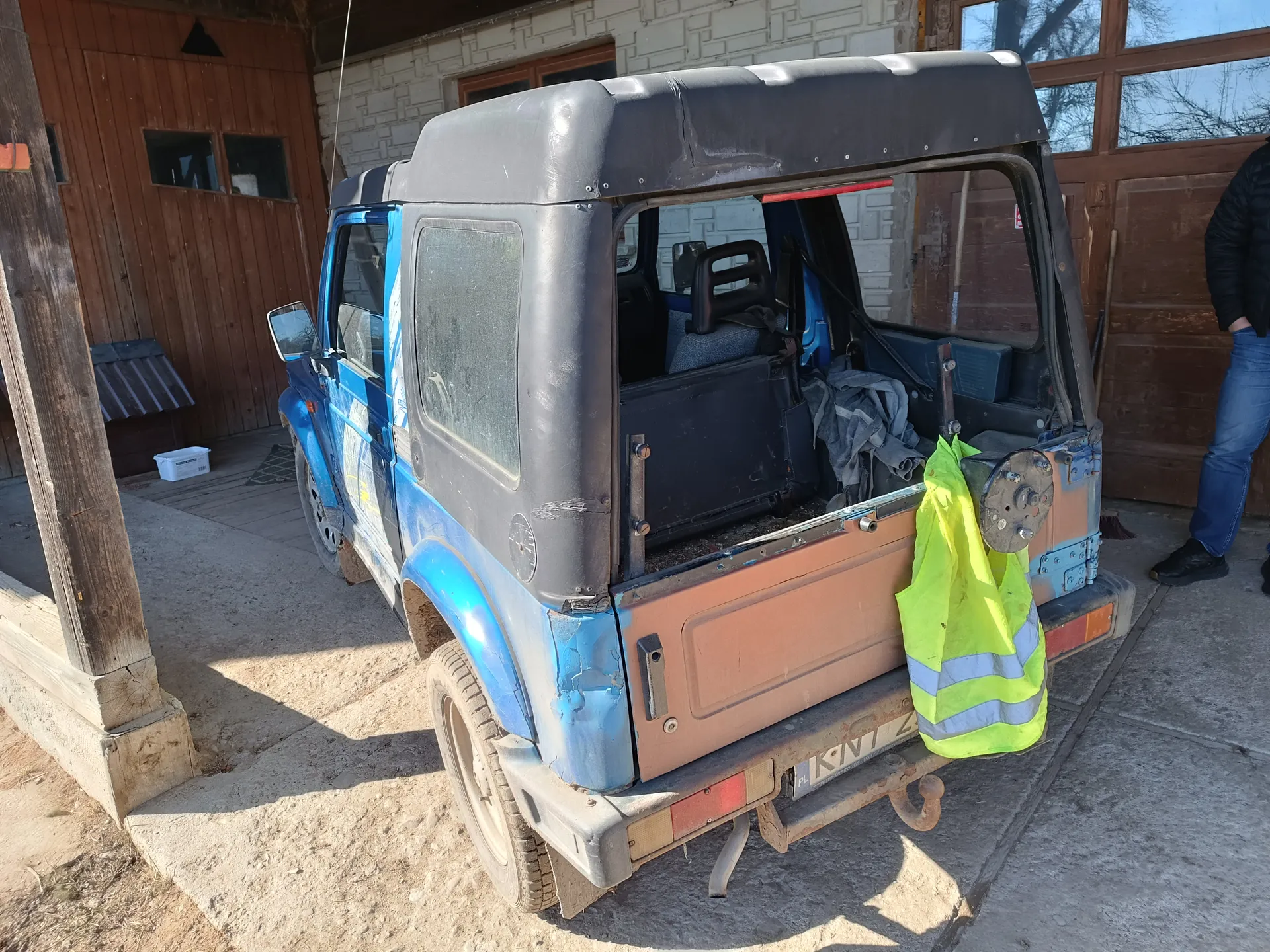 Niebiesko-czarny samochód terenowy z brązowym tyłem zaparkowany przed budynkiem z cegły i drewna. Na tylnym zderzaku wiszą żółta kamizelka odblaskowa z niebieskimi paskami oraz tablica rejestracyjna z częściowo widocznym numerem KNT 2. W tle widoczne są drewniane drzwi, okna, biały pojemnik na ziemi oraz osoba w czarnej kurtce i niebieskich dżinsach stojąca po prawej stronie. Samochód ma otwarte tylne drzwi, a wewnątrz widać siedzenia i szare materiały. Podłoga składa się z betonu i ziemi.