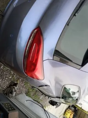Zbliżenie na tylną część srebrnego samochodu typu hatchback z widocznym czerwonym światłem tylnym. Samochód zaparkowany na podłożu z żwiru i trawy. Po prawej stronie widoczna część innego pojazdu lub konstrukcji, na tle żółty obiekt.