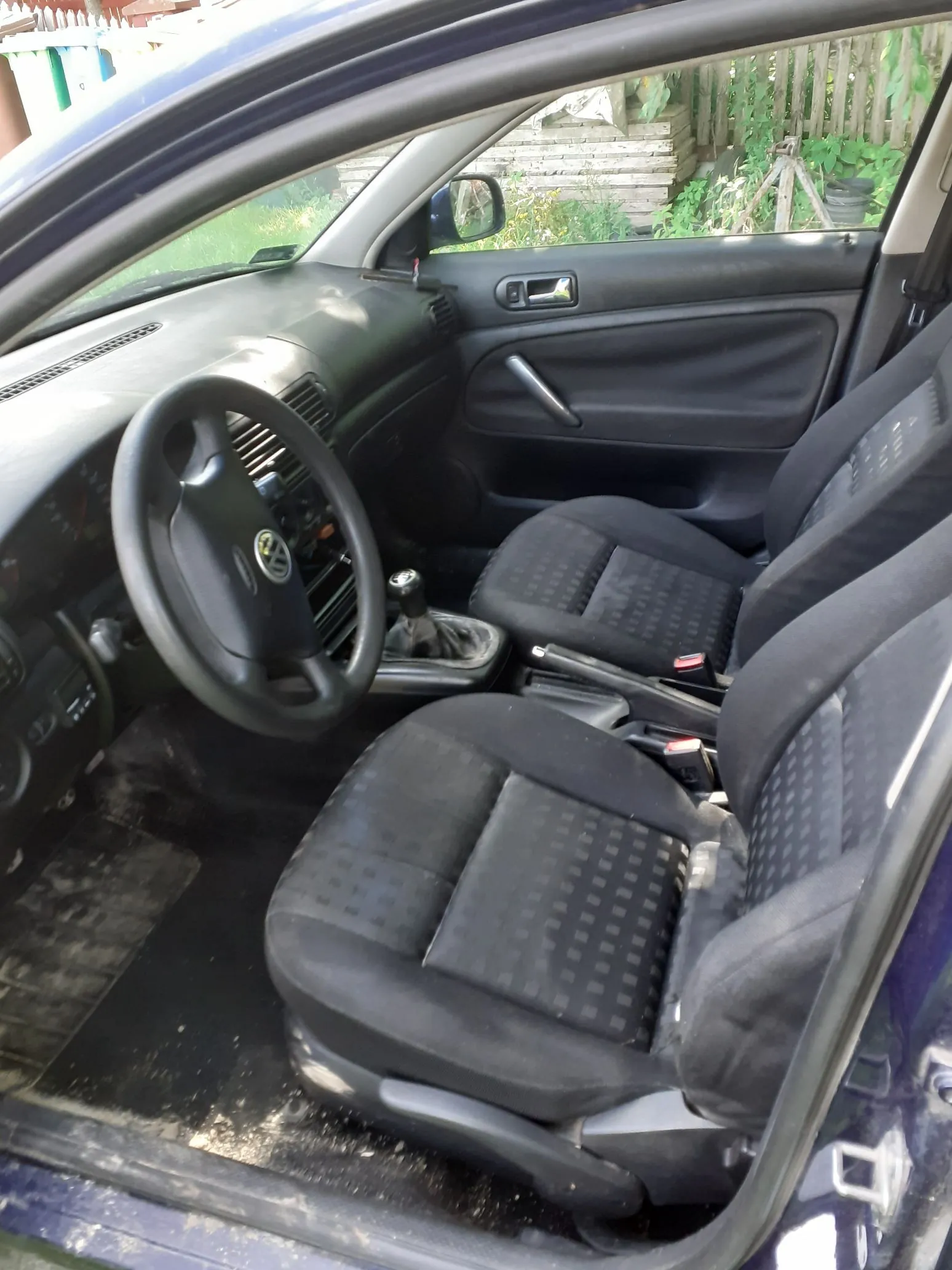 Wnętrze samochodu widziane z otwartych drzwi pasażera. Widoczna jest czarna tapicerka materiałowa na fotelach, kierownica z logo Volkswagena, deska rozdzielcza oraz panel boczny drzwi. Podłoga samochodu jest zabrudzona piaskiem i ziemią.