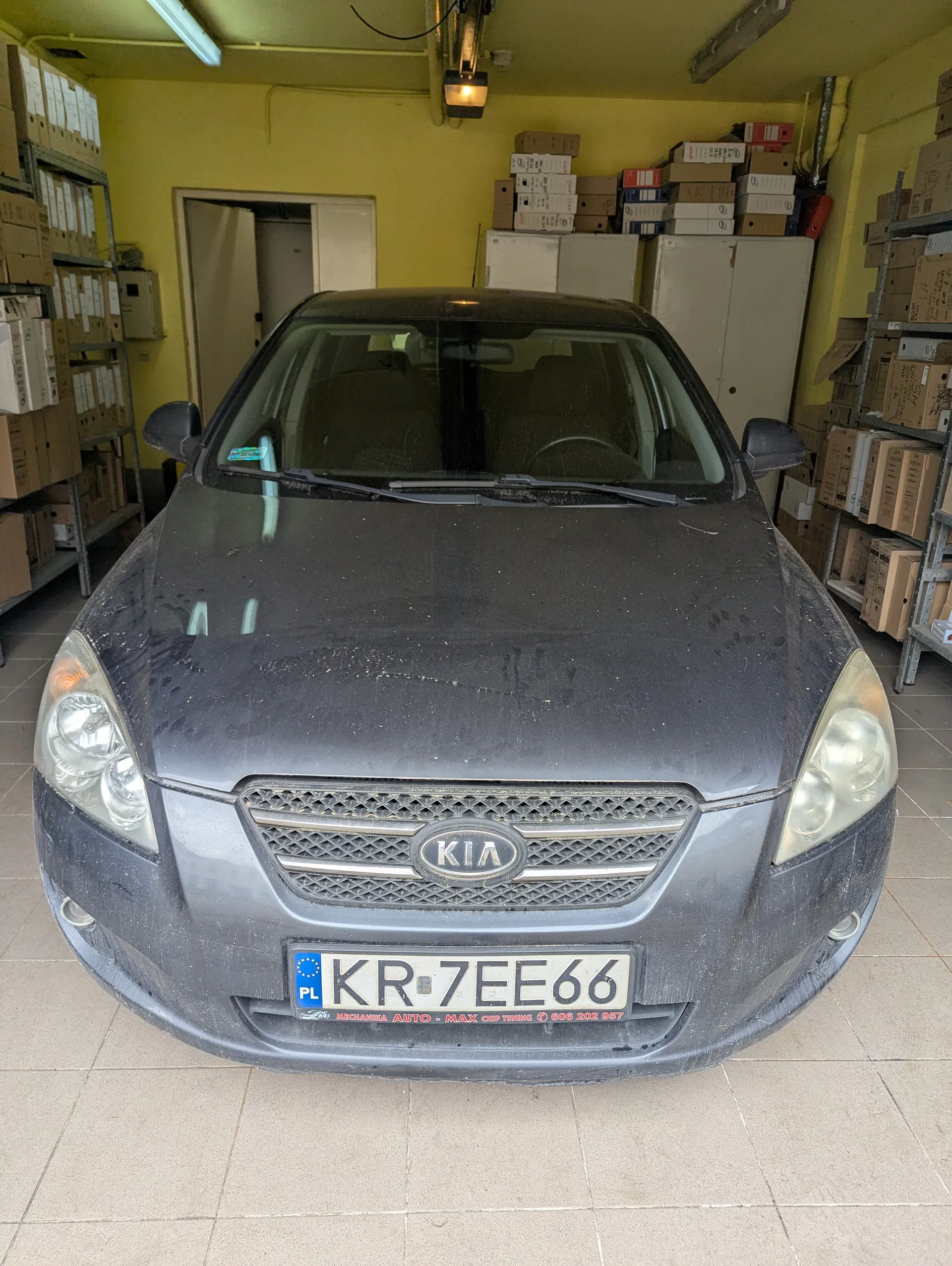 Czarny samochód marki Kia zaparkowany w pomieszczeniu magazynowym. Widoczna przód pojazdu z tablicą rejestracyjną KR-7EE66 (PL) oraz napisem "MECHANIKA AUTO - MAX CHIP TUNING" pod nią. Samochód jest zapylony. W tle żółte ściany, metalowe półki z kartonami, szafy i drzwi. Podłoga wyłożona płytkami.