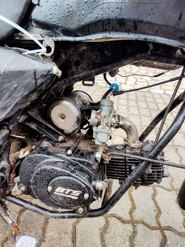 Zbliżenie na silnik motocykla z widocznymi elementami takimi jak gaźnik, przewody i blok silnika. Na bloku silnika widnieje napis "BTS". Silnik pokryty jest brudem i kroplami wody. W tle widoczna brukowana powierzchnia. Części metalowe i plastikowe są zanieczyszczone, a niektóre elementy mają ślady korozji.