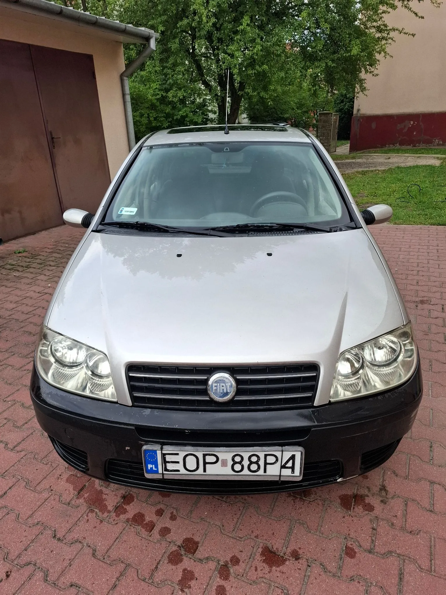 Srebrny samochód marki Fiat zaparkowany przodem do widza na czerwonej kostce brukowej. Pojazd posiada czarny zderzak przedni z polską tablicą rejestracyjną o numerze EOP 88P4. W tle widoczny jest fragment budynku z brązowymi drzwiami garażowymi oraz zielone drzewa.