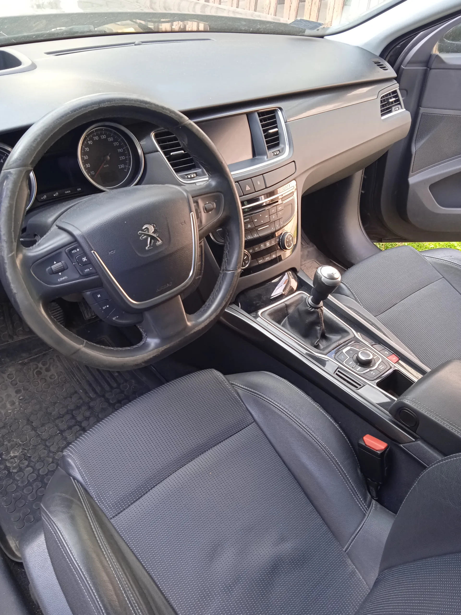 Wnętrze samochodu marki Peugeot z widocznym kierownicą z logo, tablicą przyrządów z prędkościomierzem do 230 km/h, centralną konsolą z przyciskami i dźwignią zmiany biegów, czarnymi skórzonymi fotelami z perforacją oraz otwartą drzwiami po stronie kierowcy. Na zewnątrz przez przednią szybę widoczny jest fragment ogrodzenia i trawy.