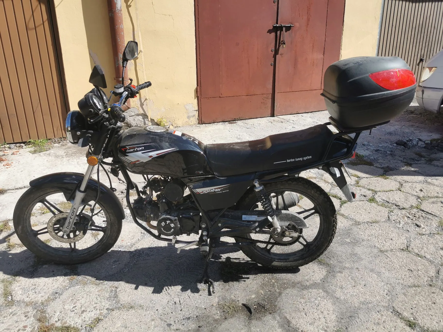 Czarny motocykl marki Barton zaparkowany na betonowym podjeździe przed budynkiem z brązowymi drzwiami garażowymi. Motocykl posiada zamontowany czarny kufer zamykany z tyłu. Na baku widnieje napis 'barton', a na boku siedzenia napis 'barton tuning system'.