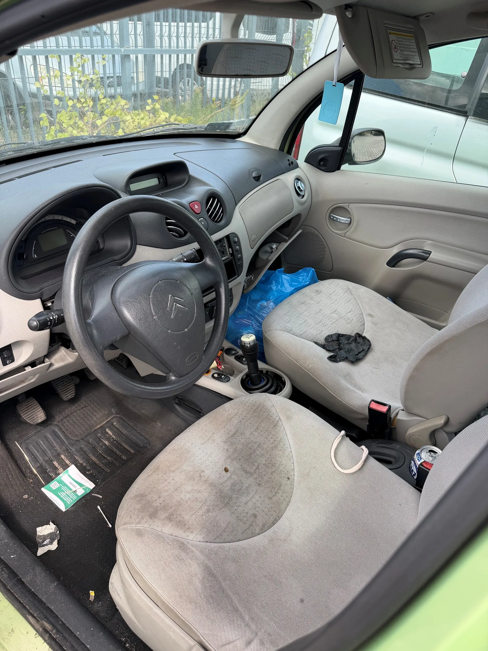 Wnętrze samochodu Citroën z widocznym kierownicą z logo marki, szarym deski rozdzielczej i beżowymi siedzeniami. Siedzenie kierowcy jest zabrudzone, na siedzeniu pasażera leży niebieska torba i czarna rękawiczka. Na podłodze widoczne są śmieci oraz zielono-biała karta. W tle przez przednią szybę widać metalowe ogrodzenie i zielone krzewy. W lusterku wstecznym widoczny niebieski znaczek.