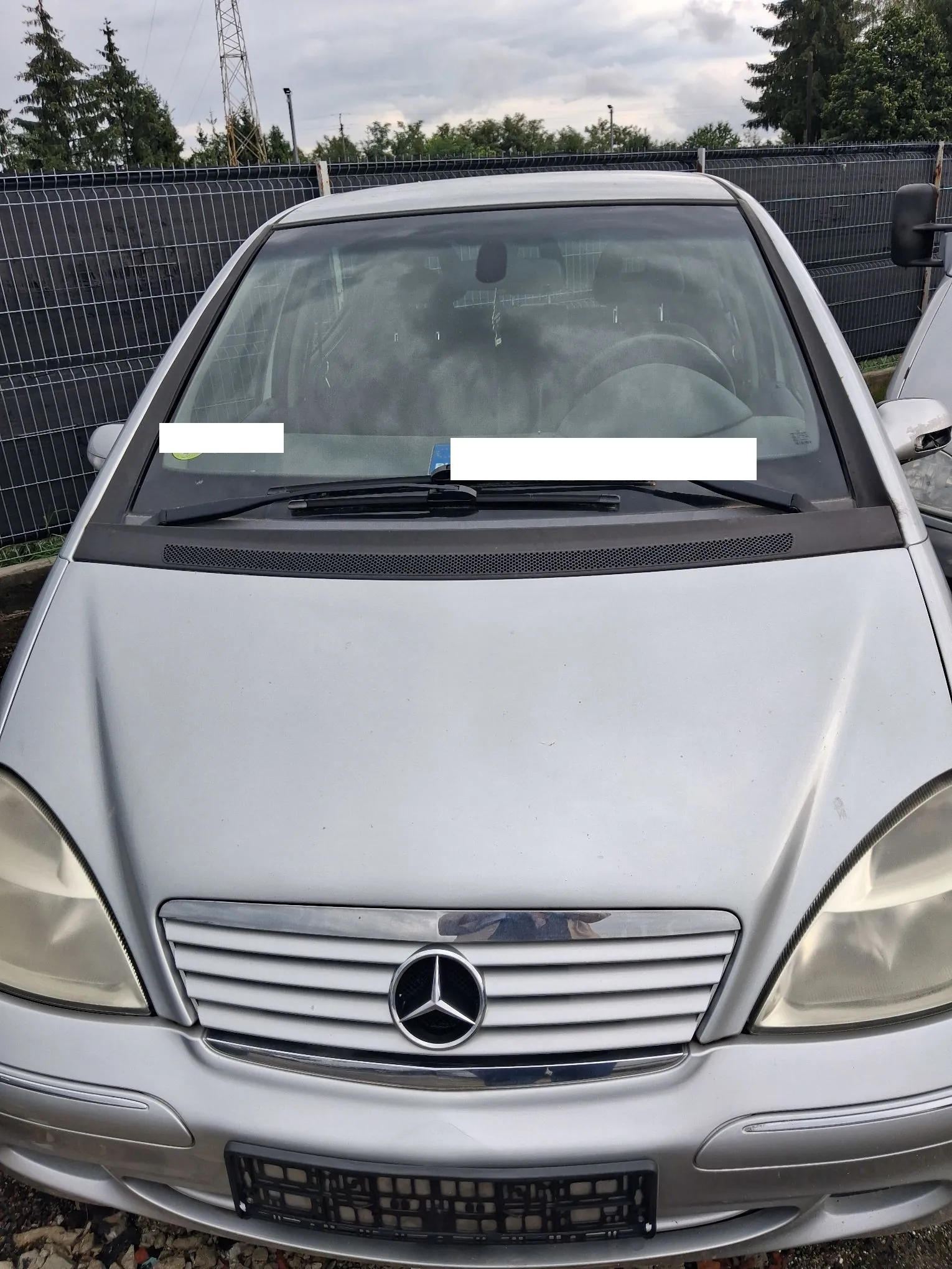 Widok z przodu srebrnego samochodu marki Mercedes-Benz. Widoczna jest maska, przednia szyba z wycieraczkami, reflektory oraz charakterystyczny grill z logo gwiazdy Mercedesa w centrum. Na dole znajduje się czarna ramka na tablicę rejestracyjną. W tle widoczny jest szary płot z siatki oraz drzewa pod zachmurzonym niebem.