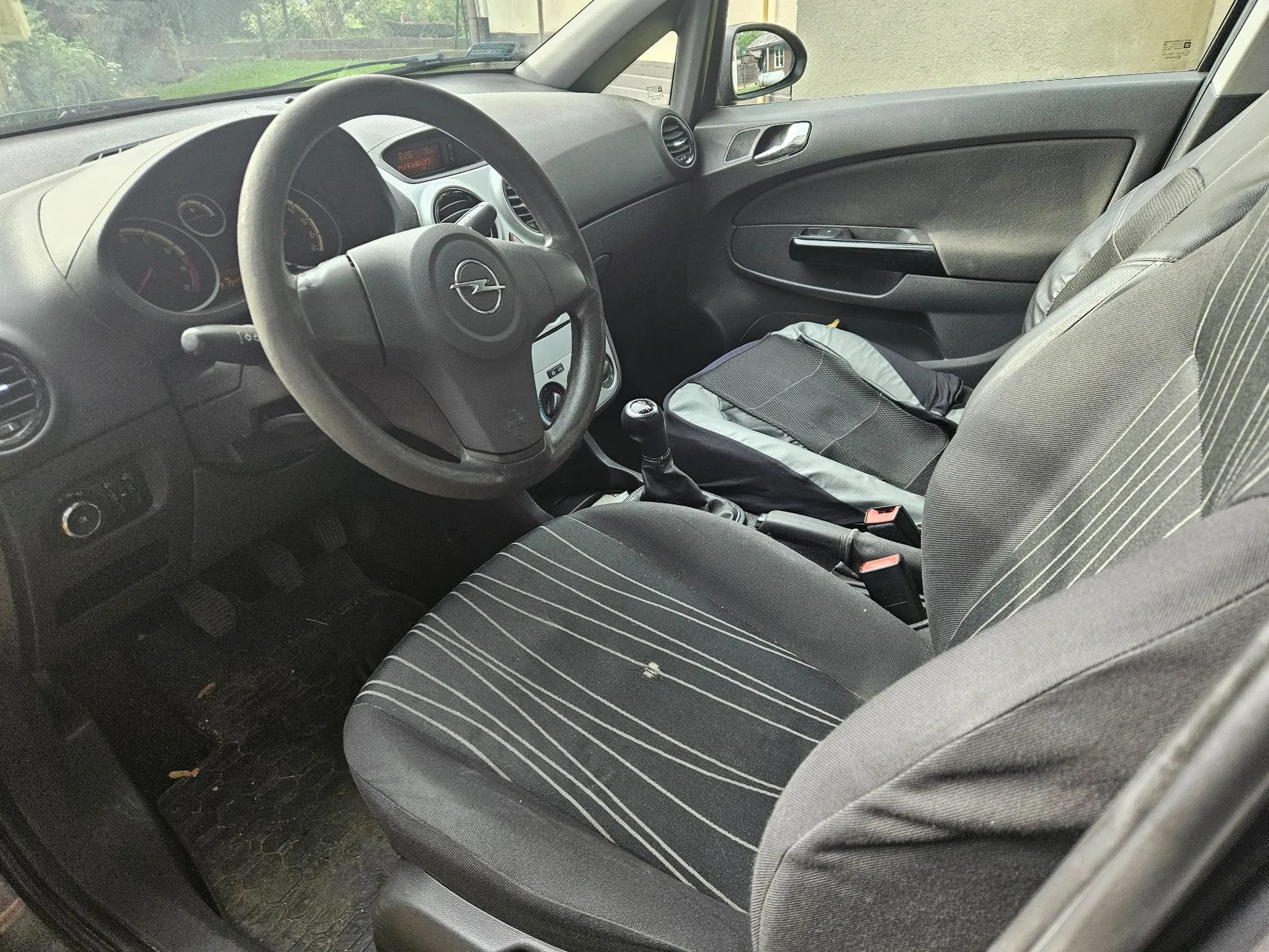Wnętrze samochodu marki Opel, widok z perspektywy pasażera na stronę kierowcy. Widoczna jest czarna kierownica z logo producenta, deska rozdzielcza z zegarami, czarne materiałowe fotele z jasnymi, pionowymi prążkami oraz przedni lewy fotel z nałożoną ciemną nakładką ochronną na siedzisku.