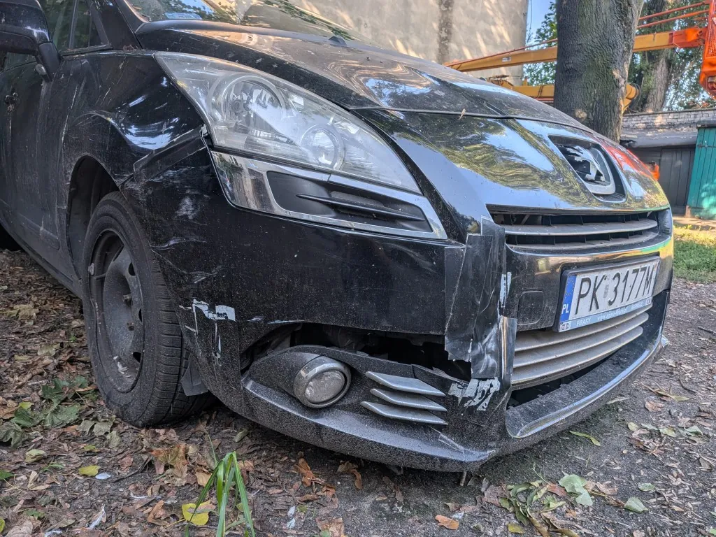Zbliżenie na przód czarnego samochodu marki Peugeot z widocznymi uszkodzeniami karoserii po prawej stronie (z perspektywy widza). Zderzak jest pęknięty i częściowo oderwany, a błotnik jest wgnieciony. Samochód ma polską tablicę rejestracyjną o numerze PK 3177M. Pojazd stoi na podłożu pokrytym suchymi liśćmi.