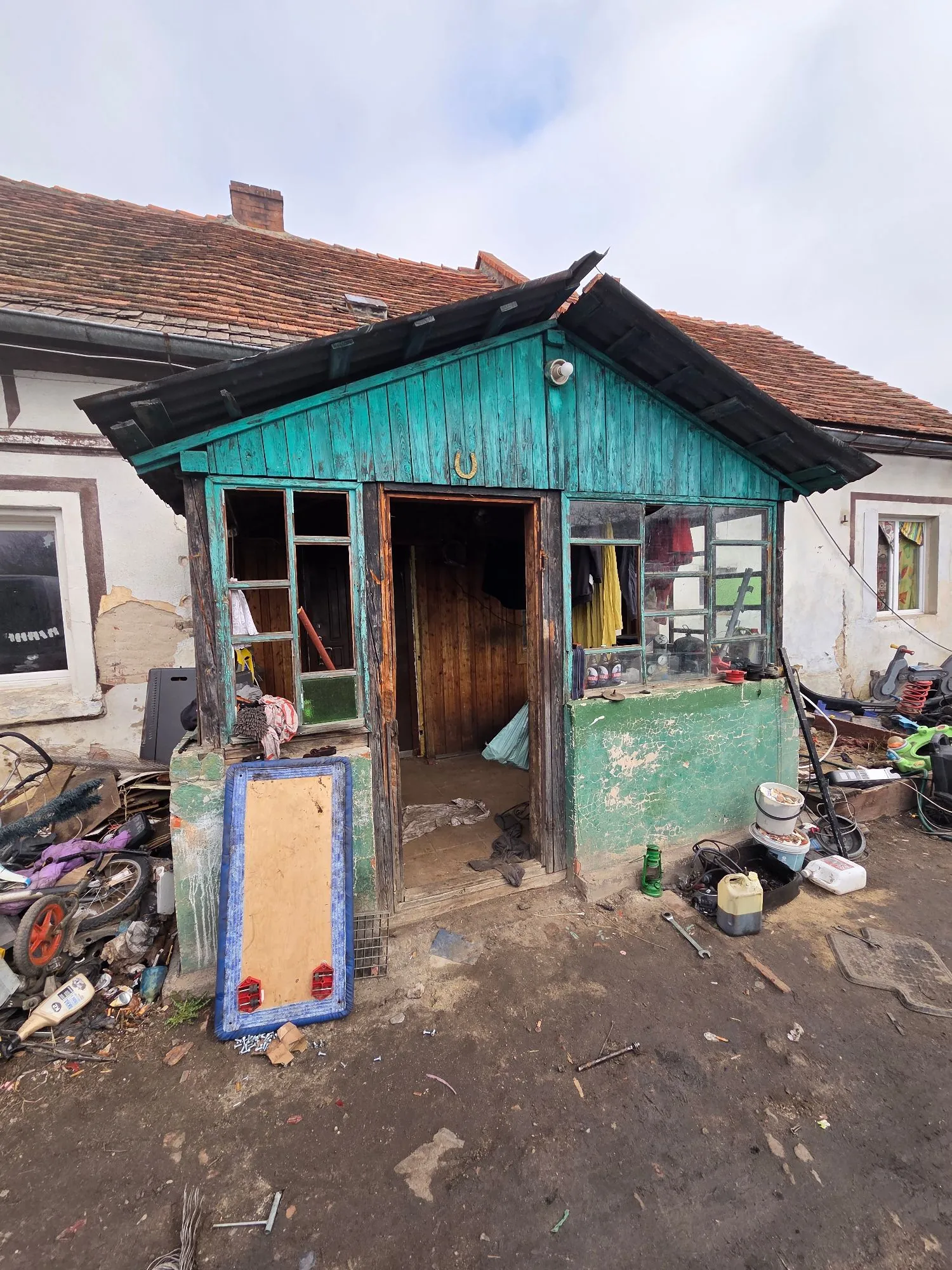 Zdjęcie przedstawia zniszczoną, drewnianą przybudówkę z turkusową farbą, dołączoną do białego budynku z brązowym, dachówkowym dachem. Przybudówka ma otwarte wejście prowadzące do ciemnego wnętrza oraz okna z drewnianymi ramami, z których niektóre są wybite. Przed budynkiem znajduje się nieuporządkowany teren z ziemi, na którym leżą liczne odpady, w tym części rowerowe, plastikowe pojemniki i kawałek płyty pilśniowej w niebieskiej ramie. Nad wejściem do przybudówki wisi mały podkowa.