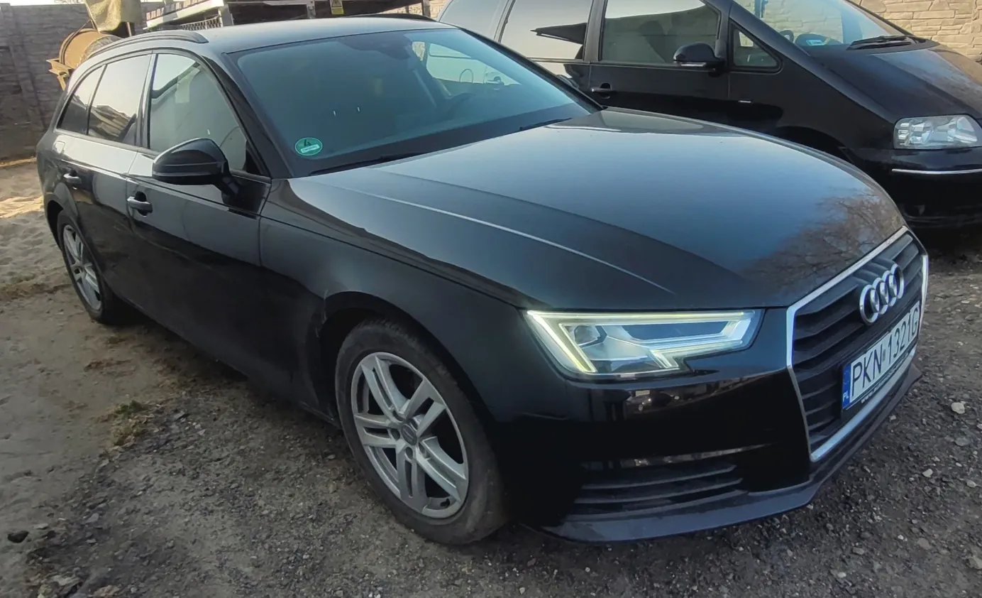 Czarny samochód Audi typu kombi zaparkowany na podłożu z żwiru. Widoczna tablica rejestracyjna PKN 1321G, logo Audi na atrapie chłodnicy oraz zielony naklejka na przedniej szybie. W tle czarny samochód osobowy i ceglana ściana. Samochód ma włączone światła przeciwmgielne.