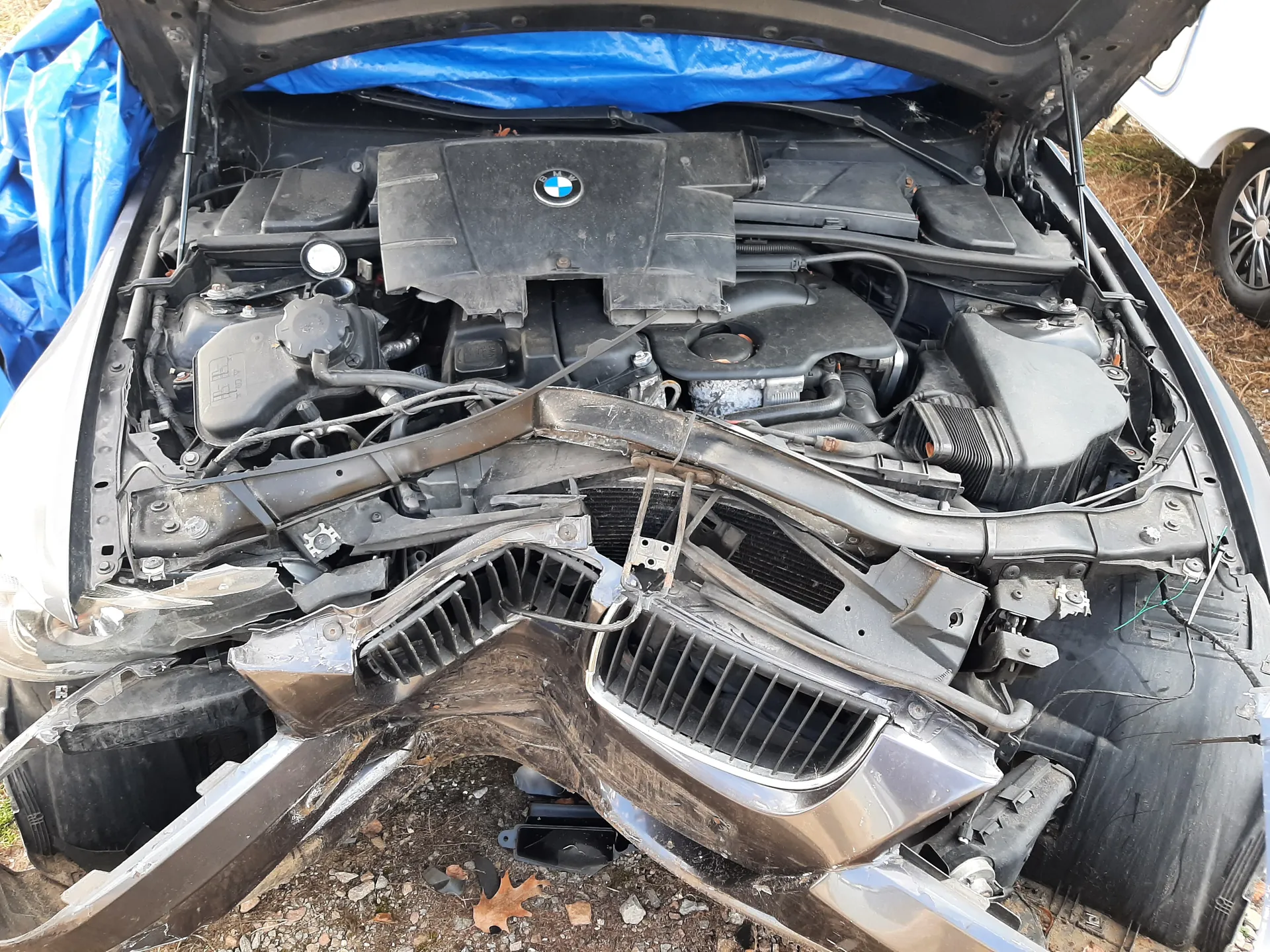 Zdjęcie przedstawia uszkodzony samochód marki BMW z otwartą maską. Widoczna jest zniszczona przód pojazdu, w tym zdeformowany zderzak i atrapa chłodnicy. W komorze silnika znajdują się elementy takie jak zbiornik płynu chłodniczego, przewody i czarna pokrywa silnika z logo BMW. W tle widoczna niebieska folia oraz fragment innego pojazdu. Podłoga pokryta jest żwirem i suchymi liśćmi.