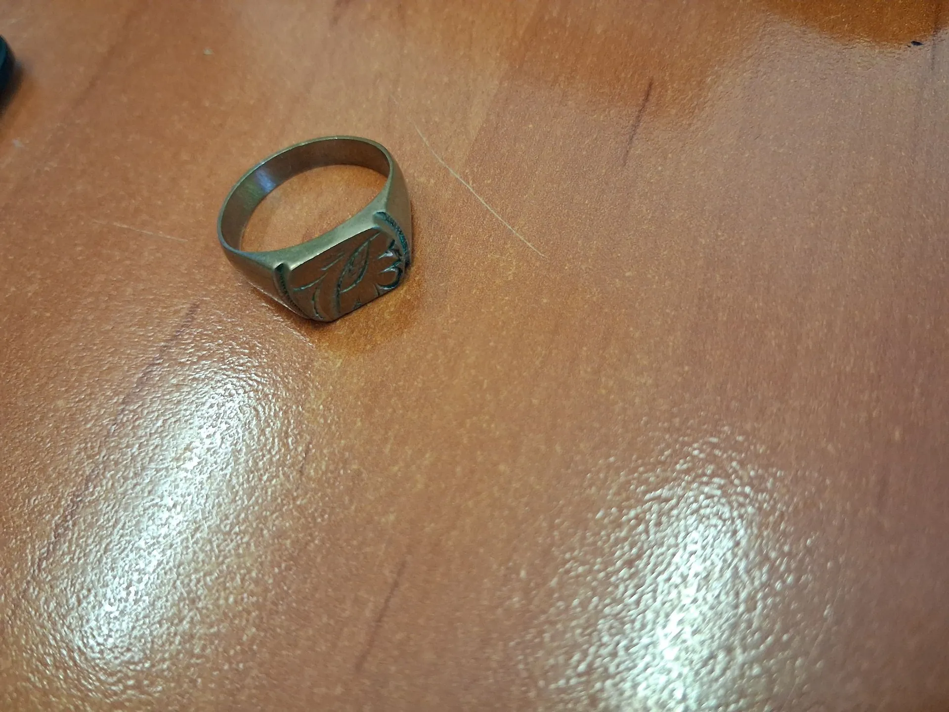 Złoty sygnet z grawerem w kształcie stylizowanego ptaka lub symbolu na płaskiej powierzchni pierścionka, leżący na brązowym, błyszczącym blacie stołu.